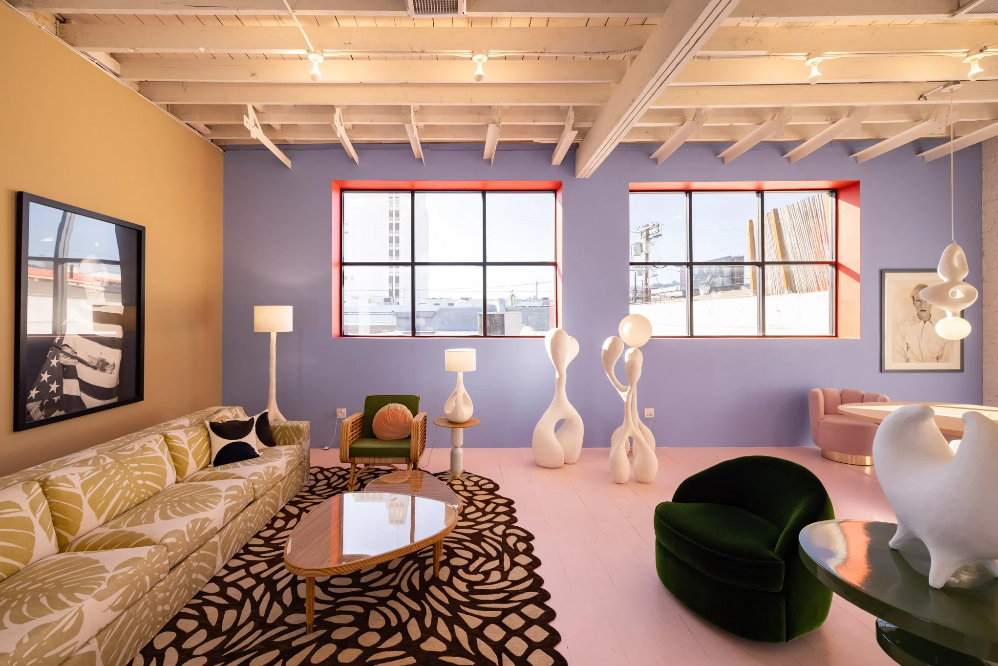 Colorful interior of a modern living room with large blue and yellow walls, large windows with red frames, a yellow patterned sofa, a green velvet chair, unique white sculptures, a black and white patterned rug, artwork on the walls, and modern light