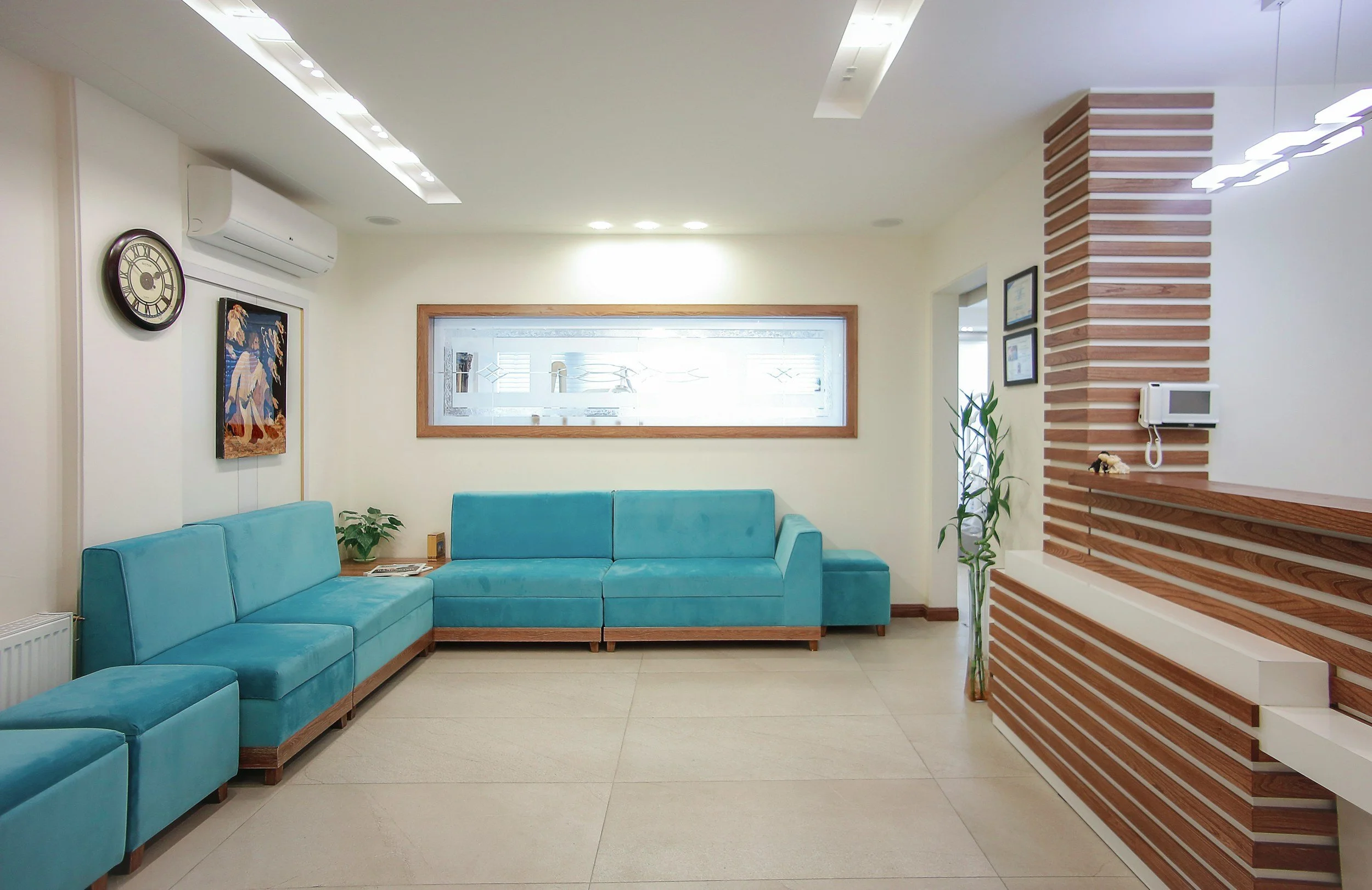 A modern living room with light-colored walls, a turquoise sectional sofa, a wall clock, a framed picture, a window with a decorative glass panel, and a wooden partition with horizontal stripes.