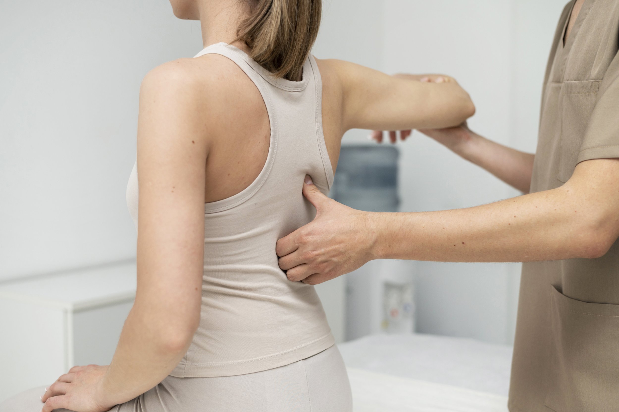 A woman wearing a beige tank top and matching pants is sitting on an examination table with her hand resting on her knee. A healthcare provider is examining her underarm with their hands.