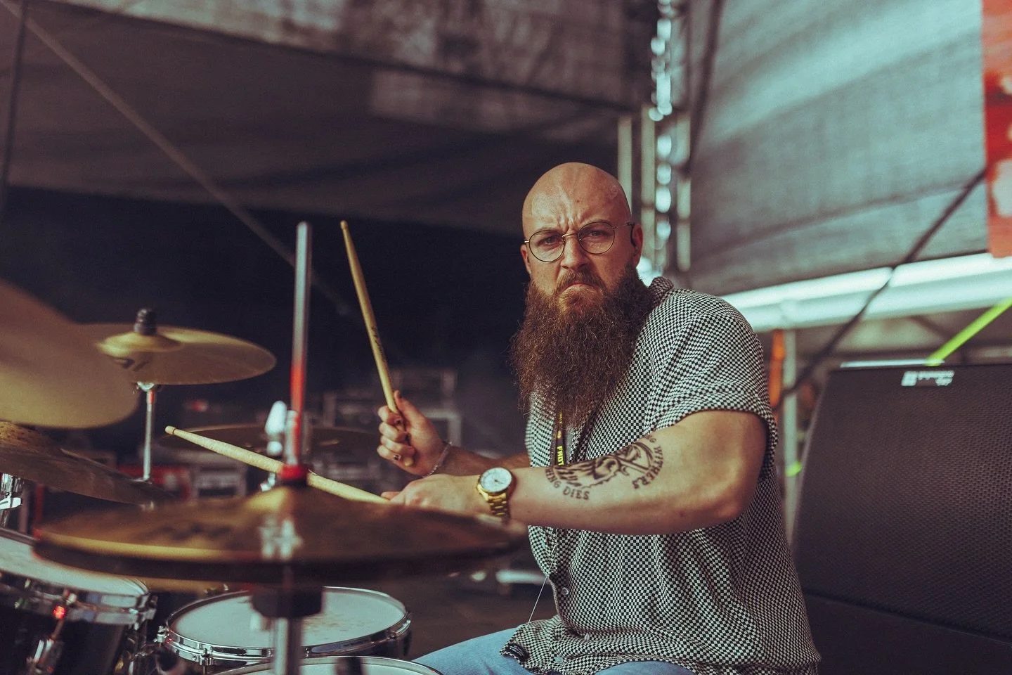 A bald man with a long beard and glasses playing drums outdoors with a serious expression.