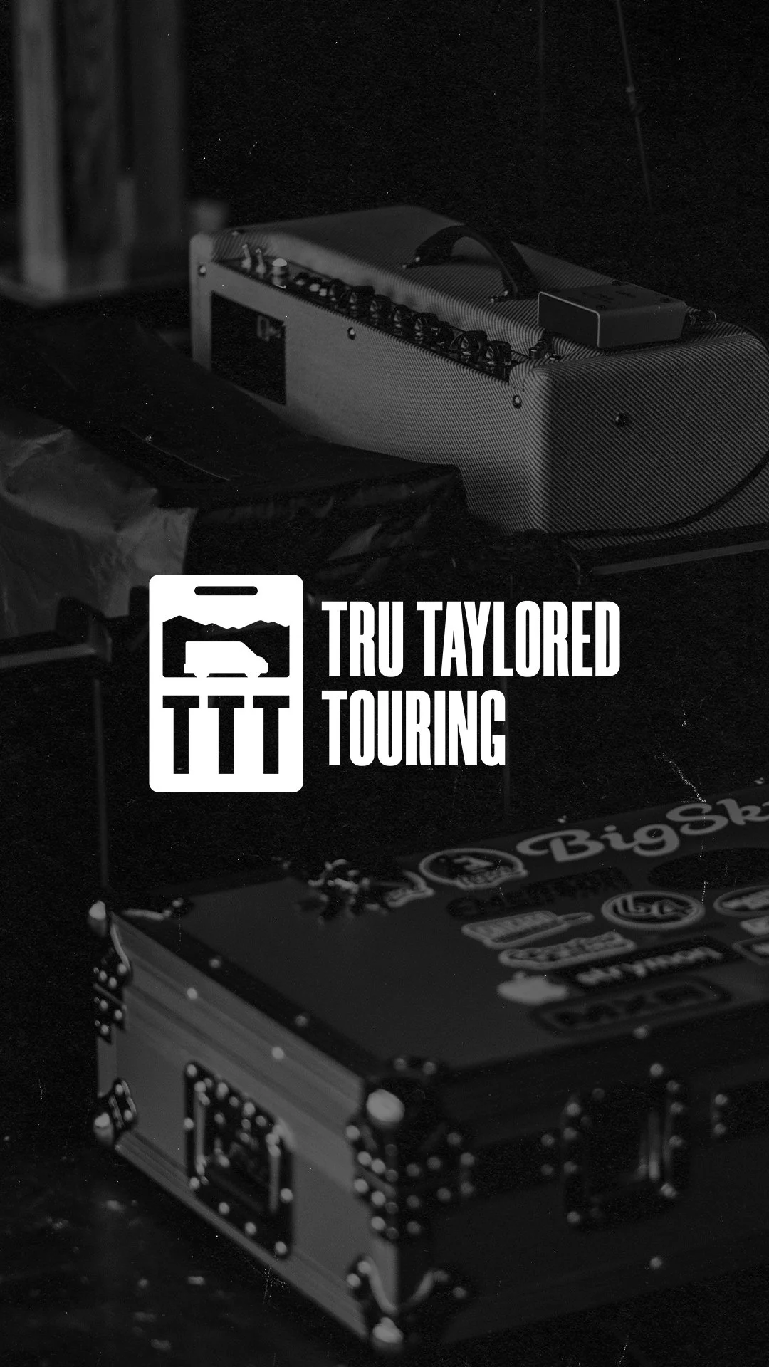 Black and white image of a guitar amplifier and a box with assorted stickers on a dark surface, with a logo and text overlay reading "TRU TAYLORED TOURING."