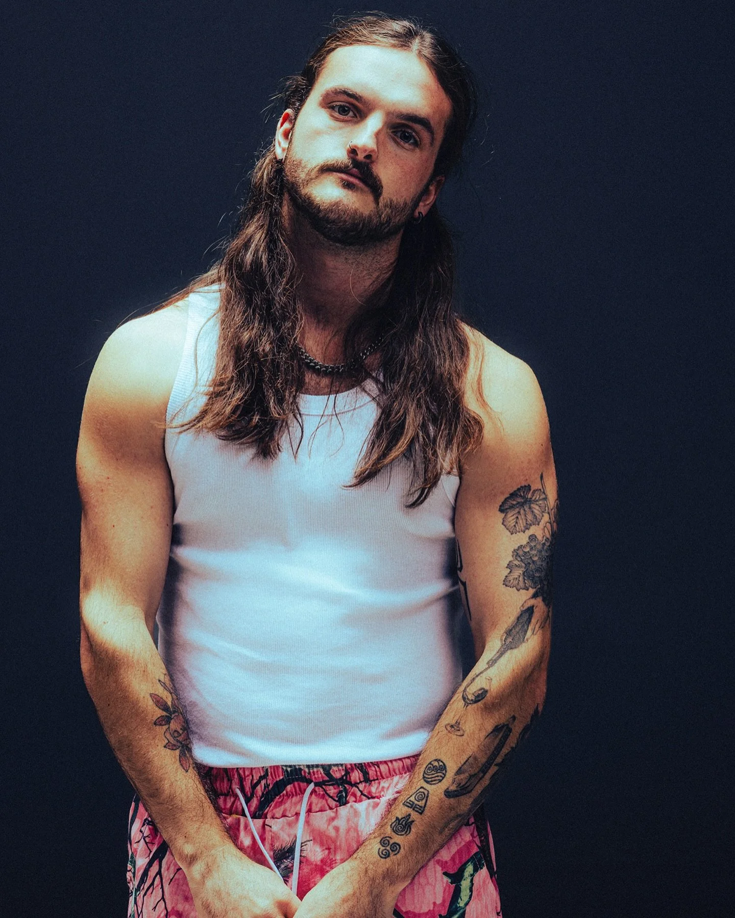 A man with long, brown hair and a short beard, wearing a white tank top and pink tie-dye shorts, standing against a dark background.