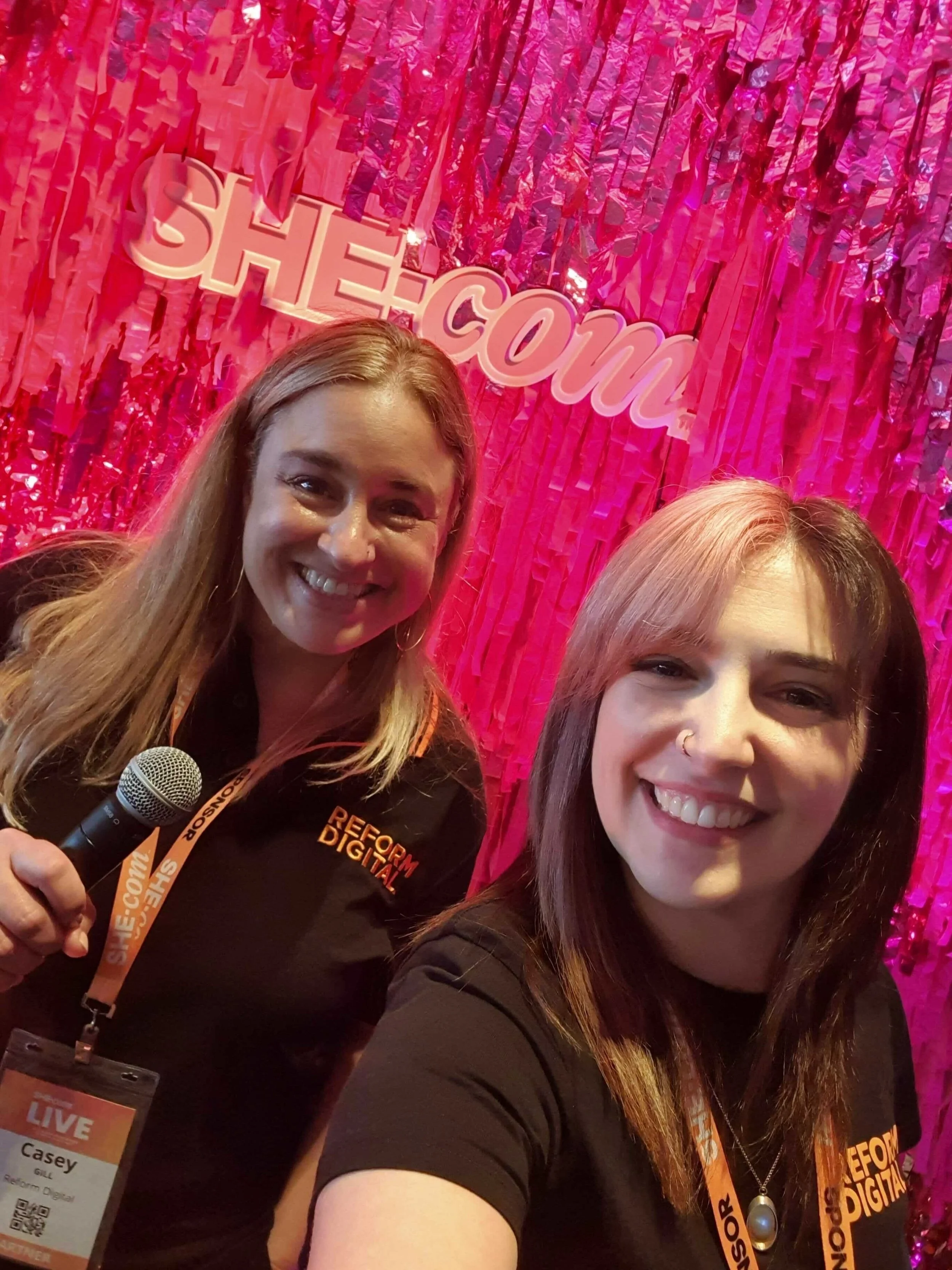 Two women smiling and taking a selfie in front of a pink, shiny backdrop with the words 'SHE.com'. One woman is holding a microphone, and both are wearing black shirts and conference badges around their necks.