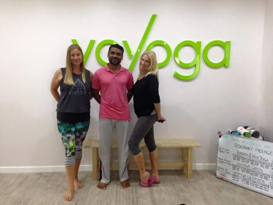 Three people standing in front of a large green 'yoga' sign on a wall inside a yoga studio, with a wooden bench and a whiteboard on the right.