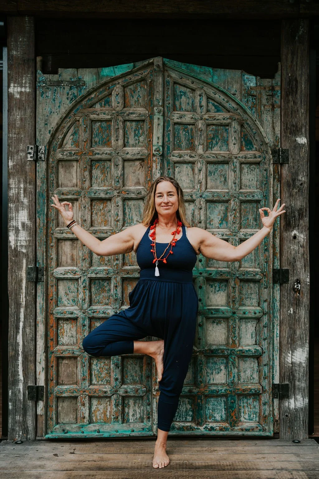 A woman practicing yoga in a tree pose with arms stretched out and one foot resting on her inner thigh, standing in front of a rustic, weathered wooden door with an arched top.