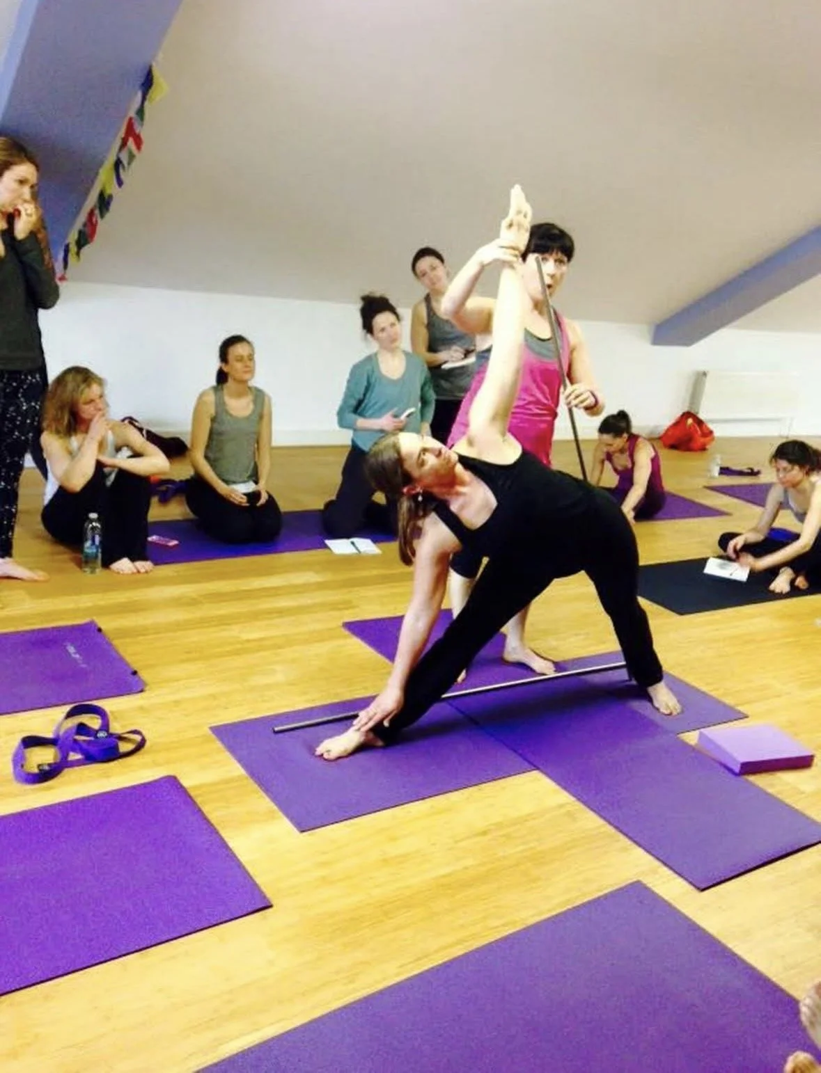 Yoga class in a studio with purple mats, one instructor demonstrating a pose while students observe and take notes.