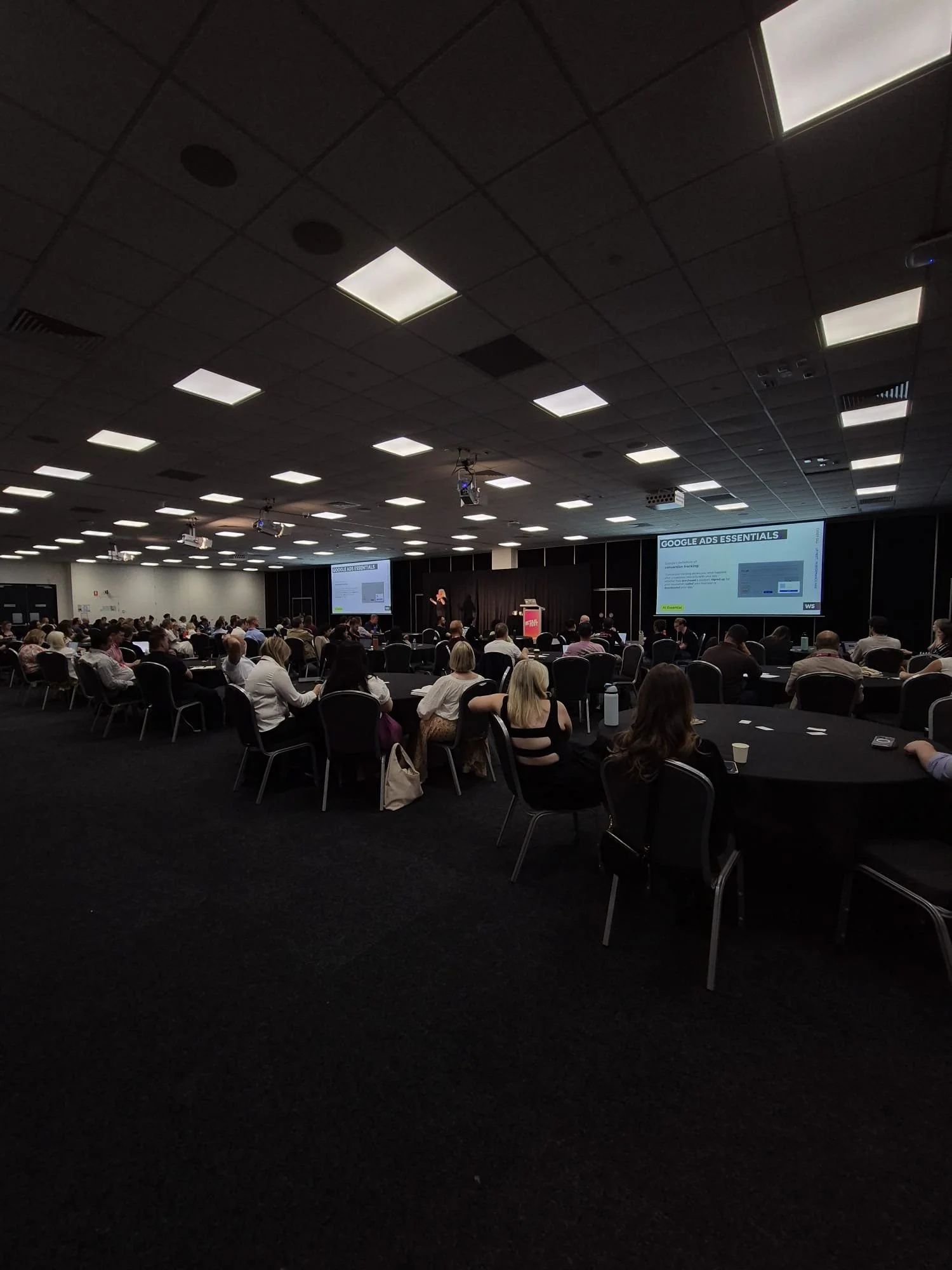 A large conference room filled with attendees sitting at round tables, facing a stage with large screens displaying a presentation about Google Ads.