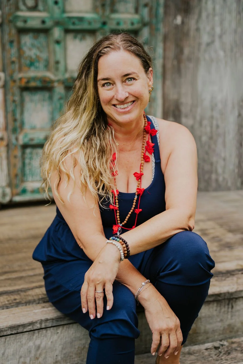 Smiling woman with long wavy blonde hair, wearing a navy blue outfit and colorful beaded jewelry, sitting on wooden steps against rustic, weathered wooden background.