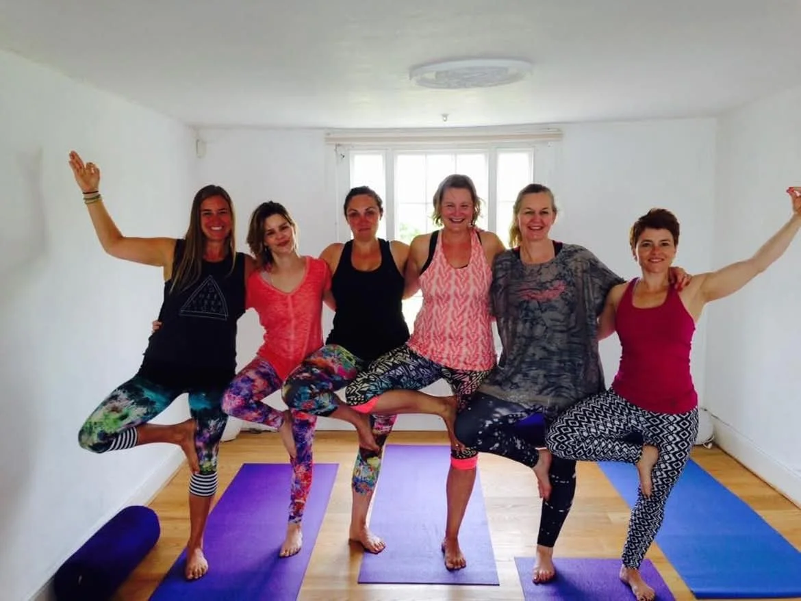 Six women doing yoga together in a bright room with windows, standing on yoga mats, and balancing on one leg with arms raised.