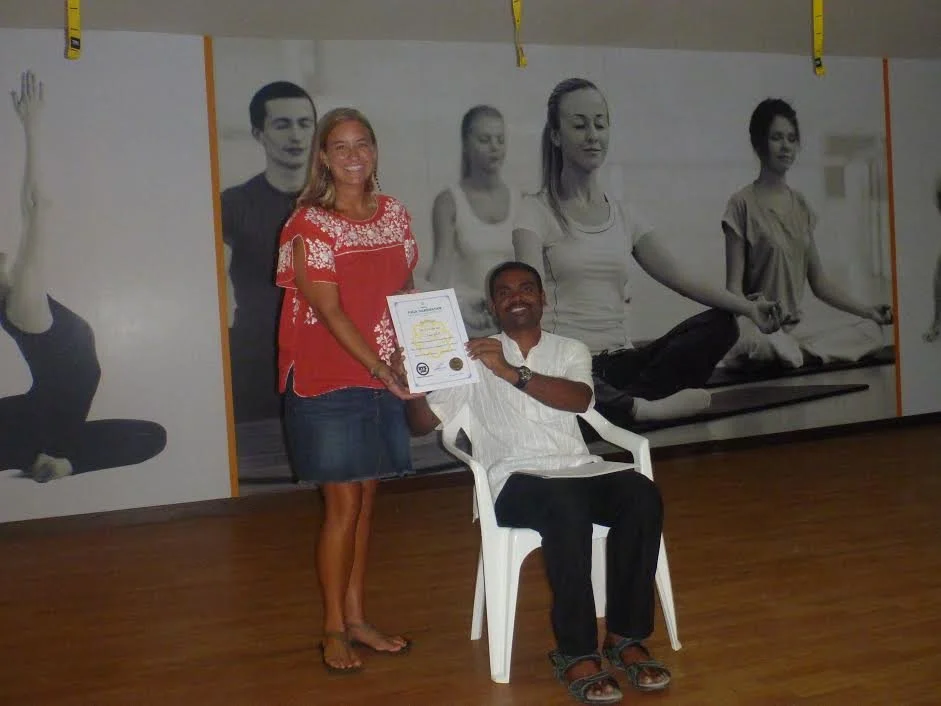 A woman in a red top and a man in a white shirt holding a certificate together, smiling. The woman stands, and the man sits on a white plastic chair in a room with bicycle-themed murals on the wall.