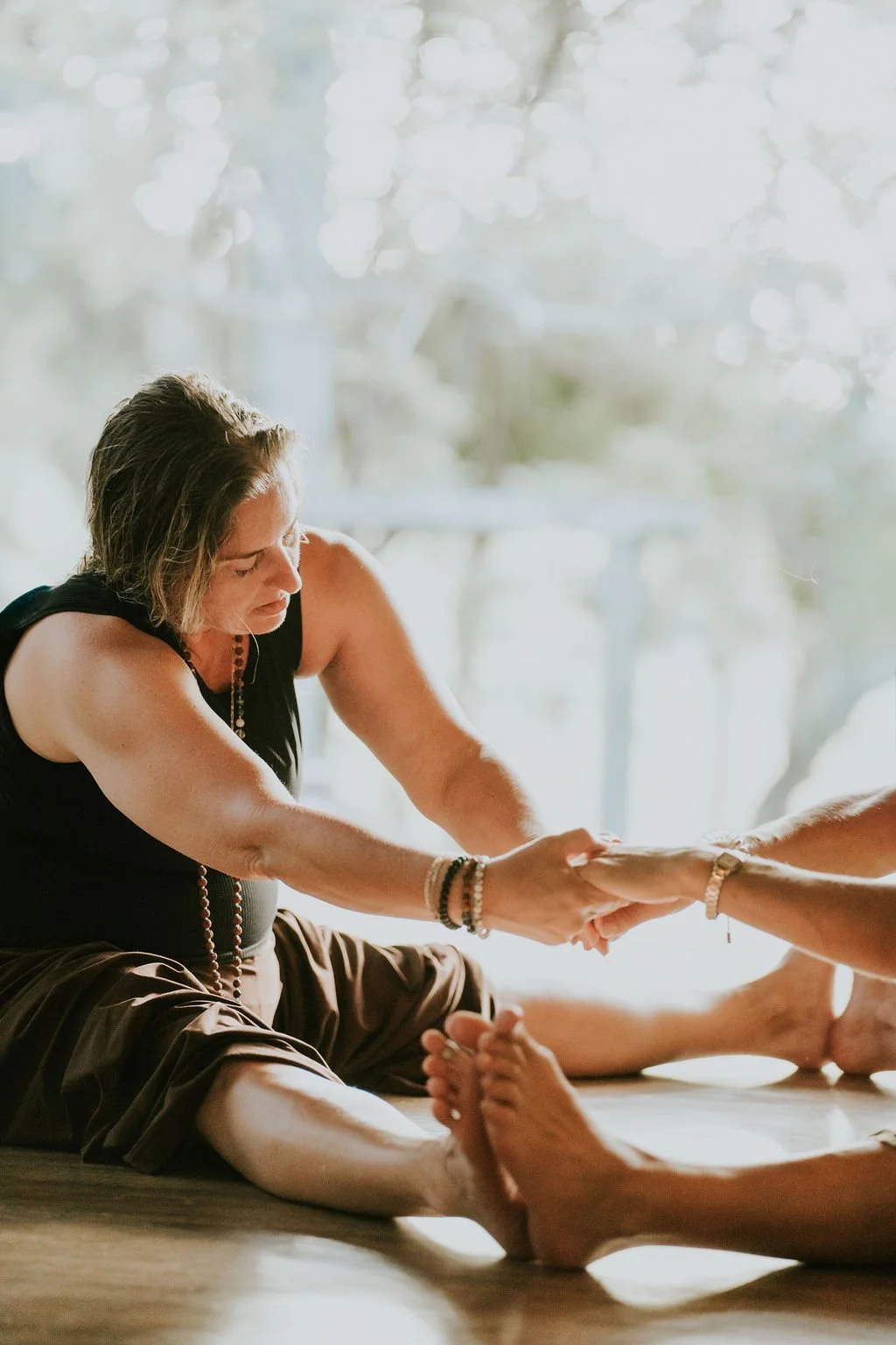 Two women practicing yoga together, sitting on the floor, holding hands, and extending their legs, with a bright and airy background featuring trees and sunlight.