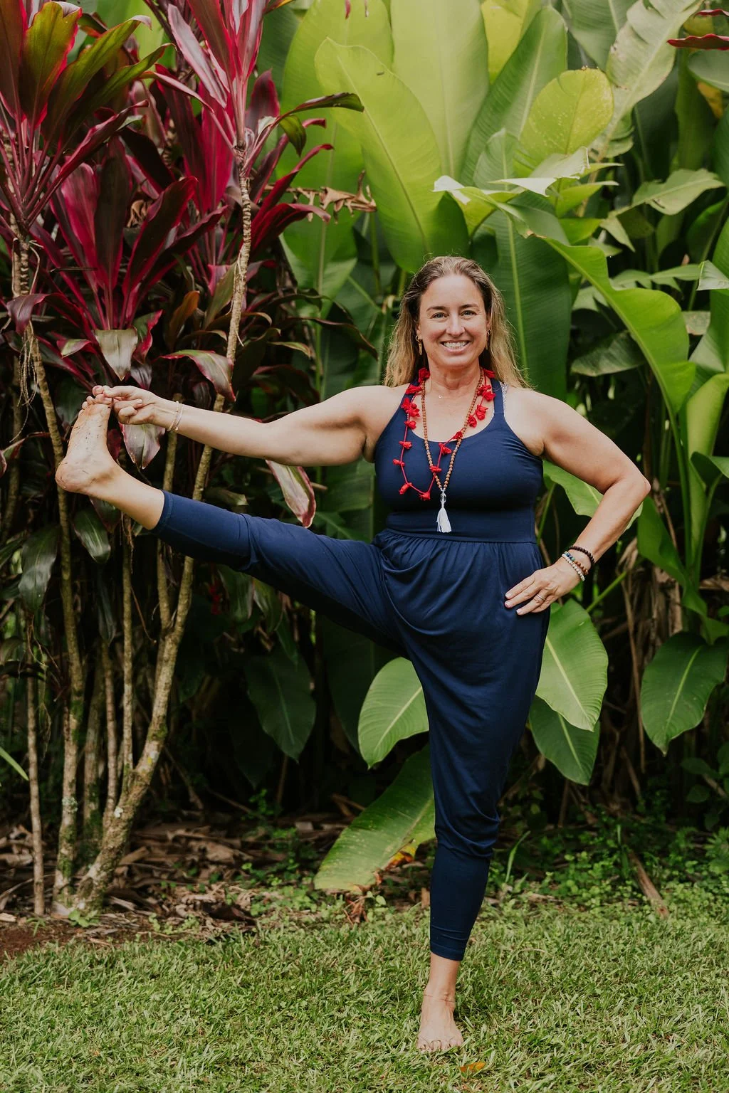 A woman practicing yoga outdoors in front of lush green and red tropical plants, standing on one leg in a yoga pose.