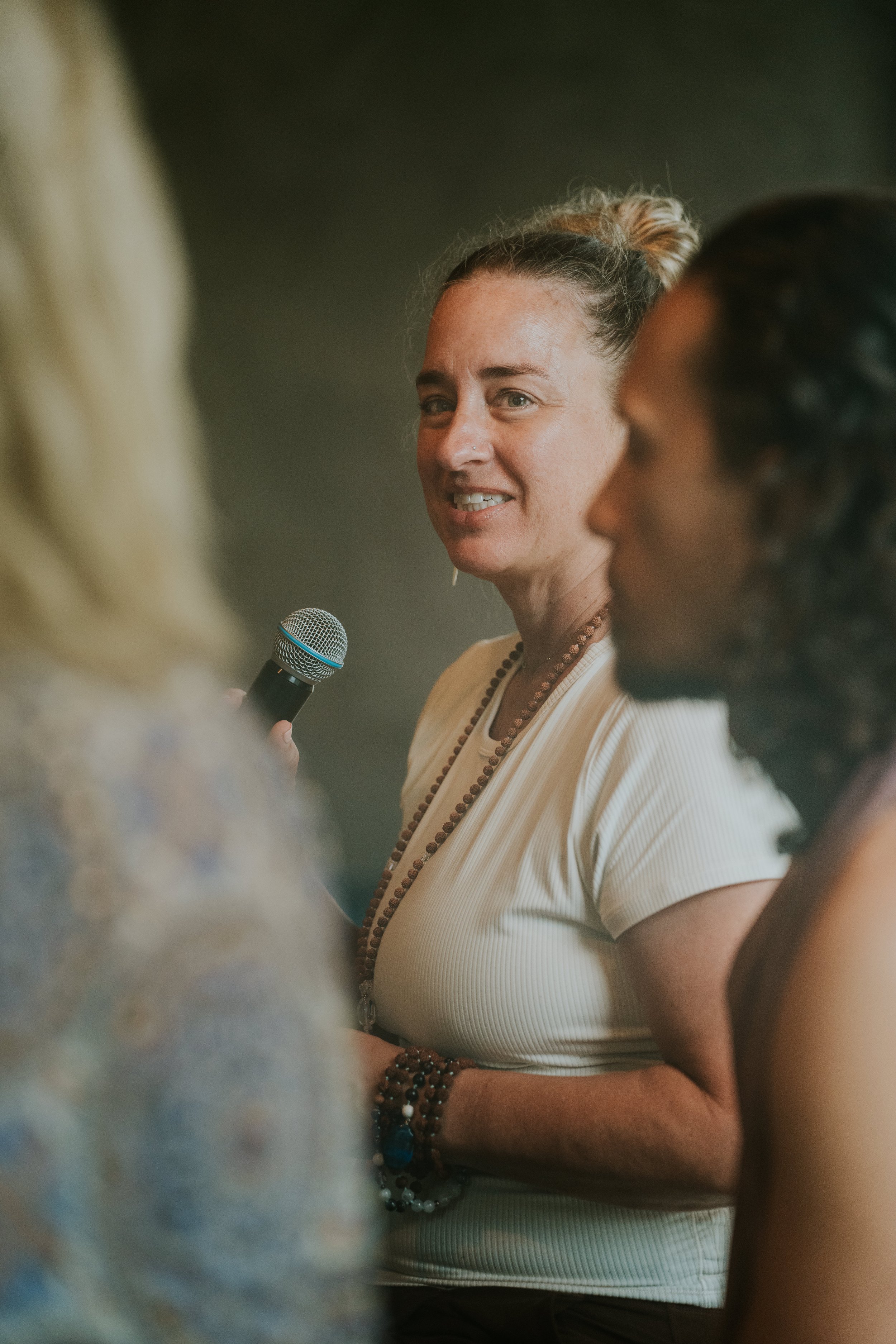 A woman with curly hair pulled back, holding a microphone, speaking to an audience. She is wearing a cream-colored shirt and beaded jewelry, and is smiling slightly.
