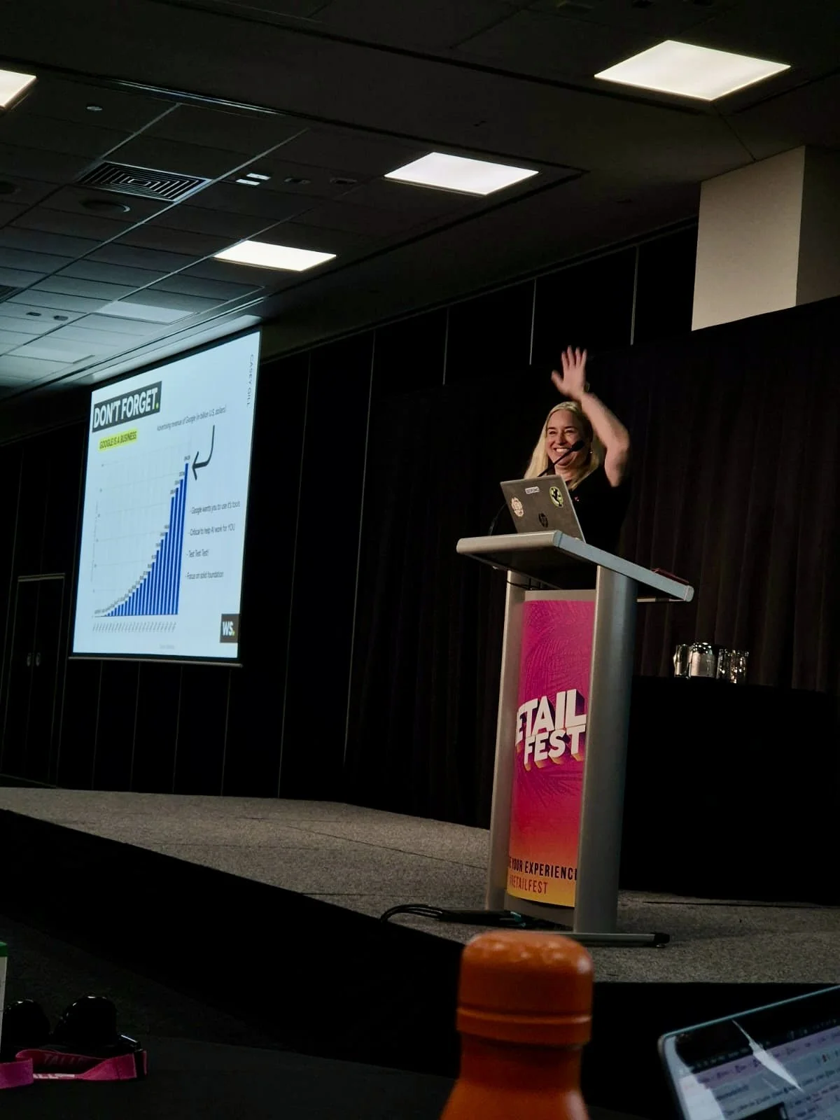 A woman with long blonde hair giving a presentation at a conference. She stands at a podium with a pink and orange banner. Behind her, a large screen displays a bar graph and the words 'DON'T FORGET' The woman is smiling and waving to the audience.