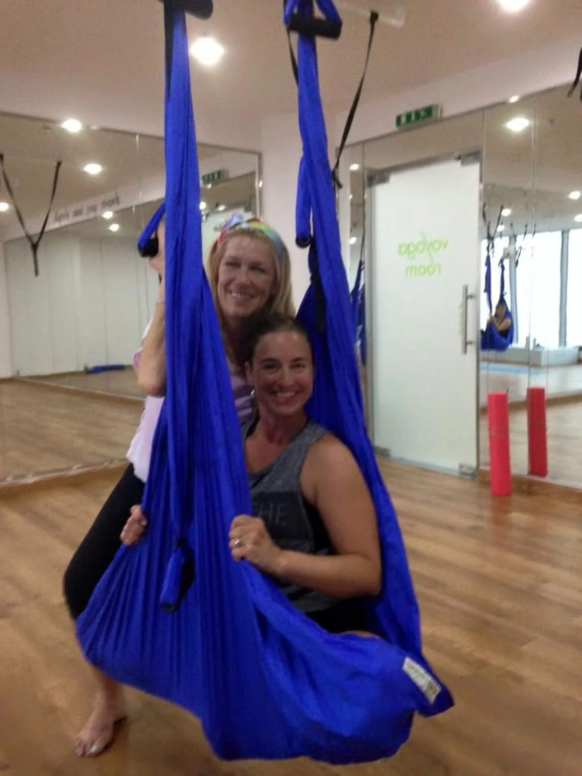 Two women smiling in a fitness studio, with one sitting in a blue aerial hammock and the other standing behind her, both holding onto the hammock.