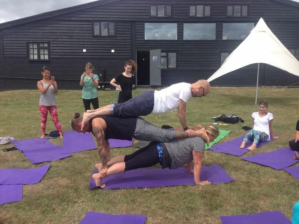 Group of people participating in outdoor yoga or acro yoga session on purple mats, with some standing and taking photos or videos, and three individuals performing a stacked yoga pose in front of a black building and a white tent.