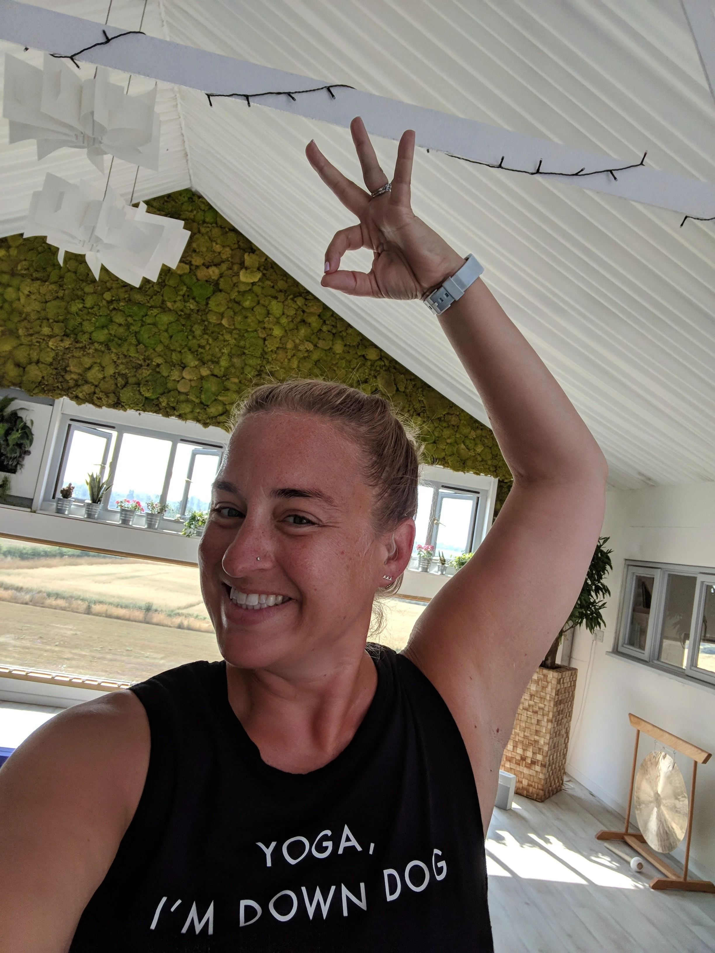 A woman smiling and raising her arm with her hand making an 'OK' gesture while taking a selfie inside a bright room with decorative elements, including a moss wall, a large window, and a harp.