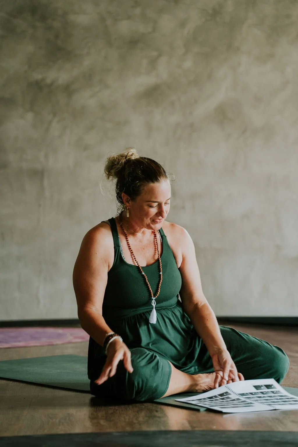 A woman practicing yoga indoors, sitting cross-legged on a yoga mat, looking at a printed black-and-white photos or images.