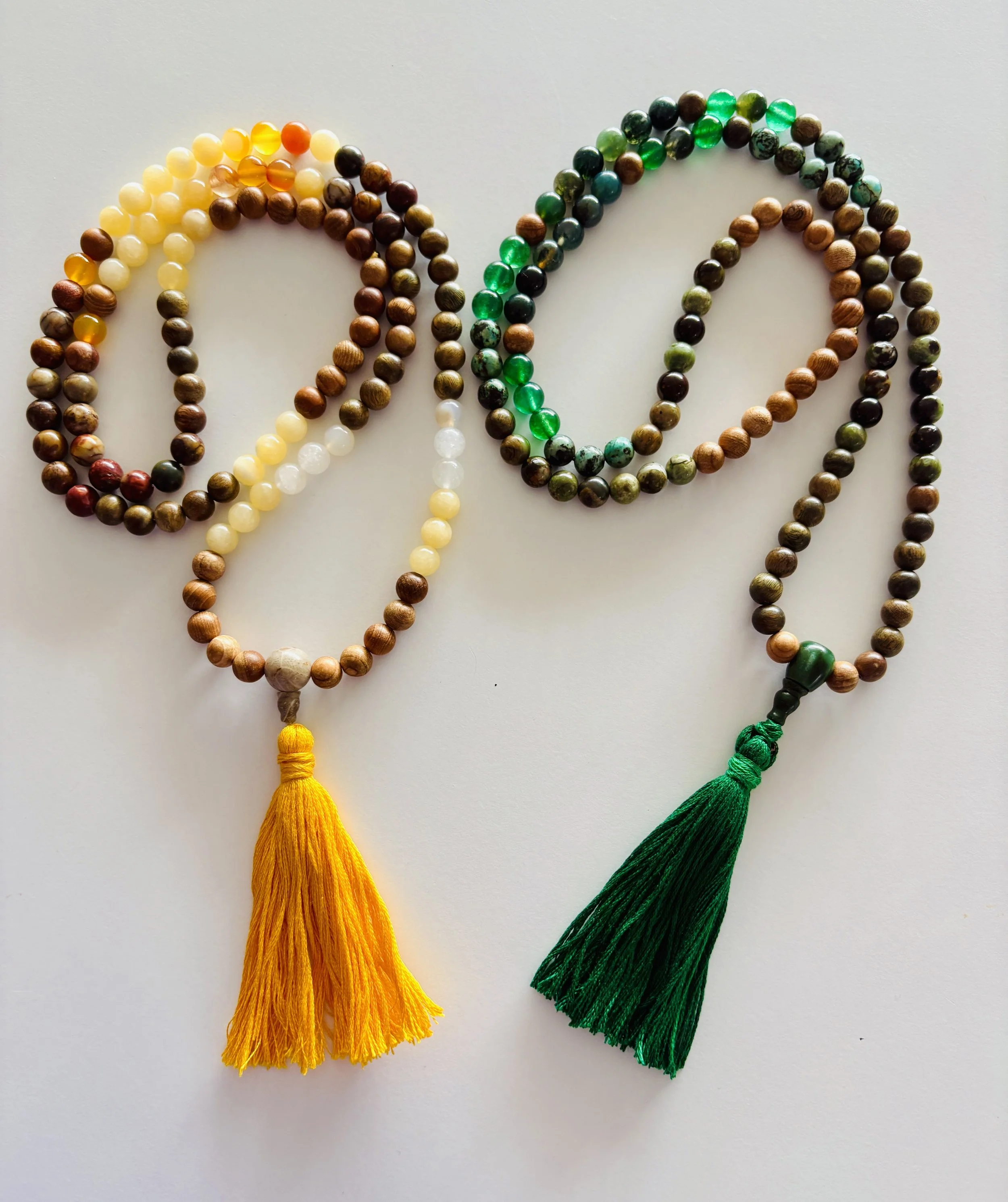 Two beaded prayer malas with tassels, one with yellow tassel and one with green tassel, arranged on a white surface.