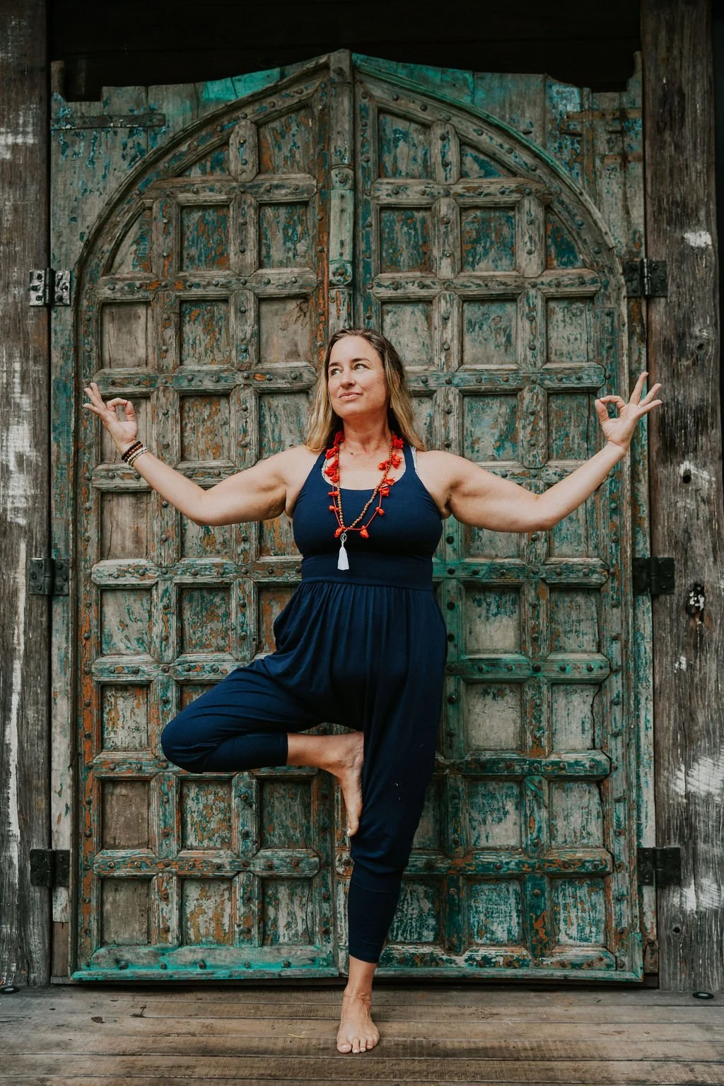 A woman practicing yoga in a tree pose with her eyes closed, wearing a navy jumpsuit, standing on one foot, and holding her other foot with her hand. She is performing in front of an ancient, weathered wooden door with turquoise accents.