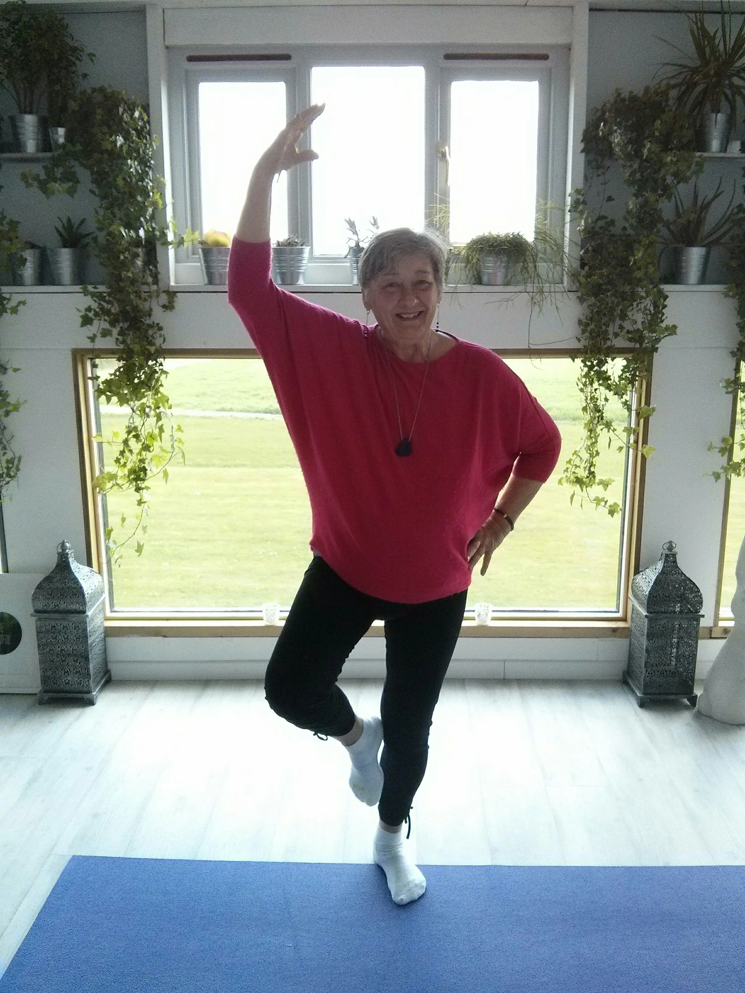 A smiling elderly woman in a pink top and black pants standing on a yoga mat in a room with large windows and plants, striking a yoga pose with one arm raised and one foot balanced on the other ankle.