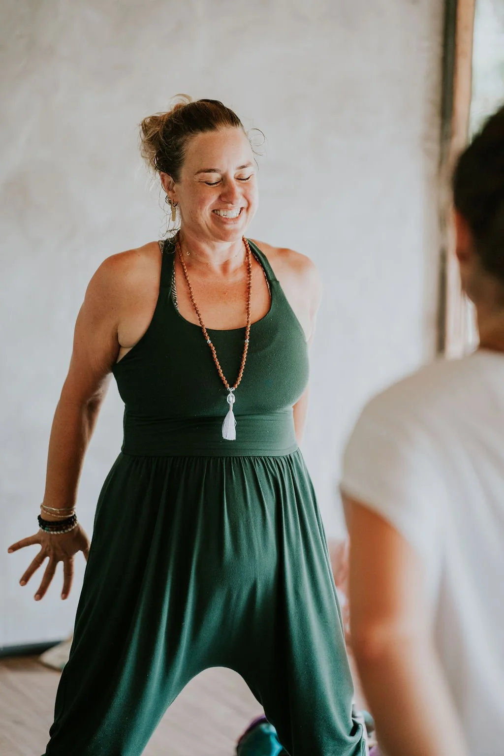 Woman smiling and standing with hands behind back, wearing a green tank top and pants, with beaded jewelry, in a room with natural light.
