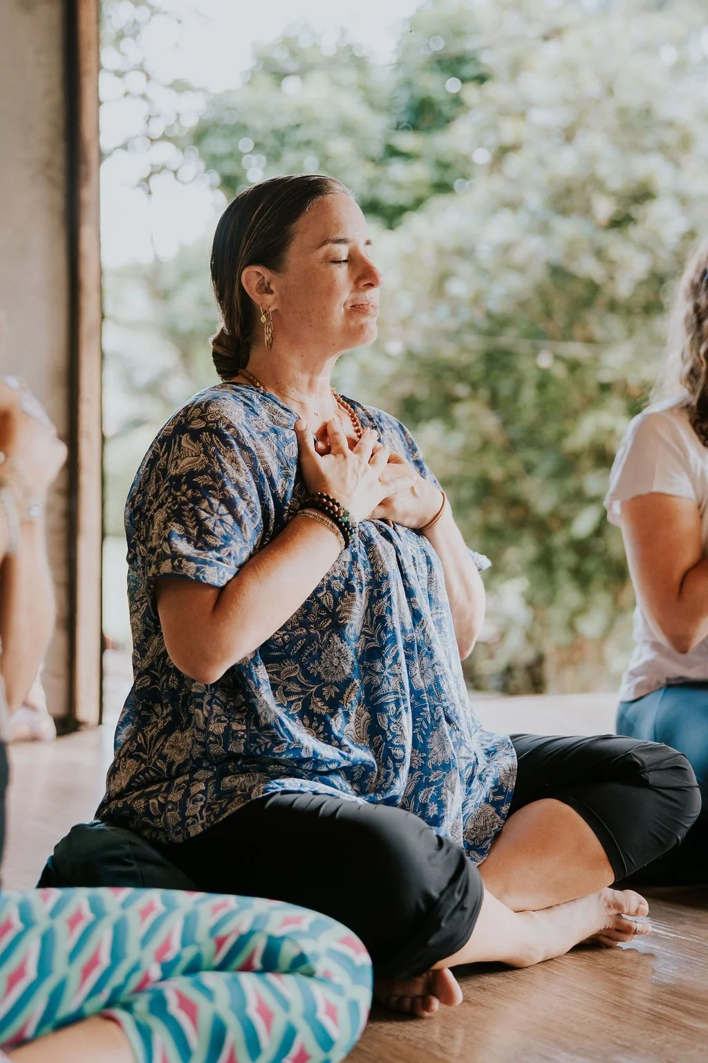 A woman sitting cross-legged on the floor with her eyes closed and hands on her chest, engaging in a meditation or mindfulness practice next to a window with trees outside.
