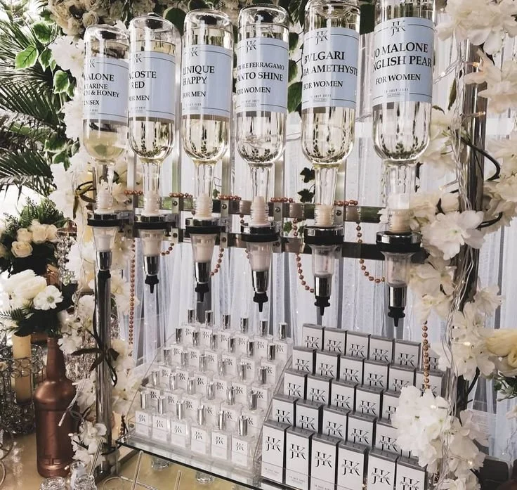 A display of luxury perfume bottles with spray nozzles, arranged on a stand decorated with white flowers. Each bottle has a label indicating different perfume names like 'LONE MARINE', 'UNIQUE HAPPY', 'AFTER SHINE', 'Bvlgari Amethyst', and 'DOLCE & GABBANA ENGLISH PEARL FOR WOMEN'. There is a collection of smaller boxed perfumes or related items below the bottles.