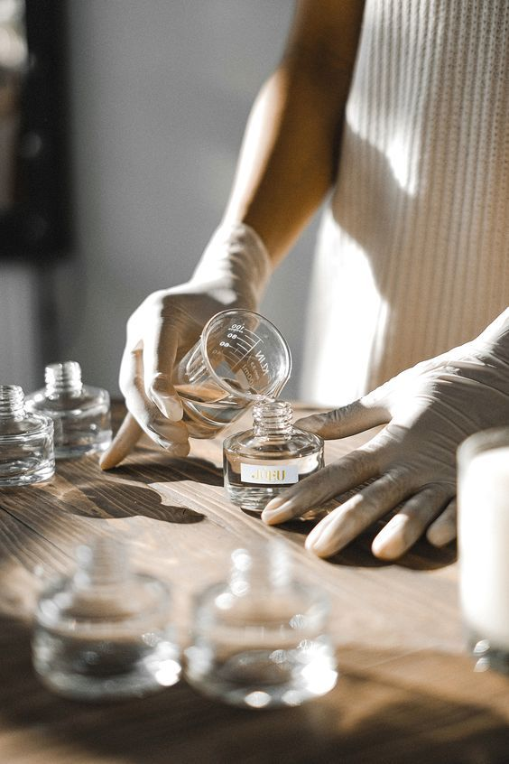 Person pouring a liquid into a small glass perfume bottle on a wooden table, wearing white gloves.