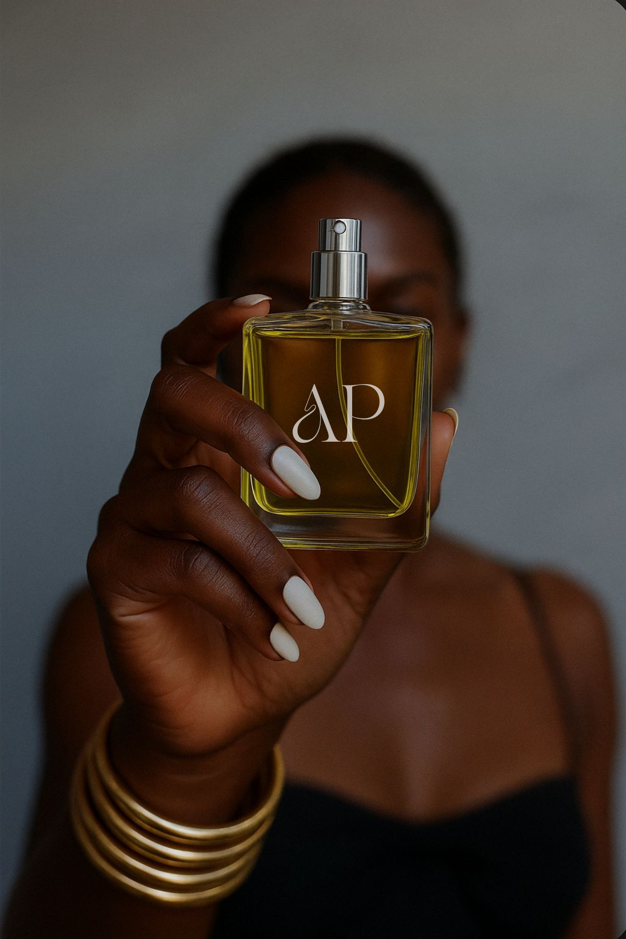 A person with dark skin and white painted nails is holding a clear perfume bottle with yellow liquid and white lettering close to the camera. The person is wearing gold bangles and a black top, with a blurred background.