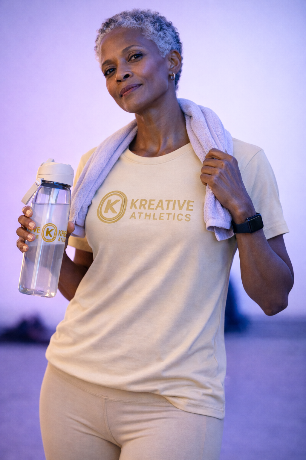 A confident older woman with short, curly gray hair stands holding a clear water bottle with a beige lid and a logo, carrying a towel on her shoulder, and wearing a beige T-shirt with the words 'Kreative Athletics' printed in gold, against a purple-l