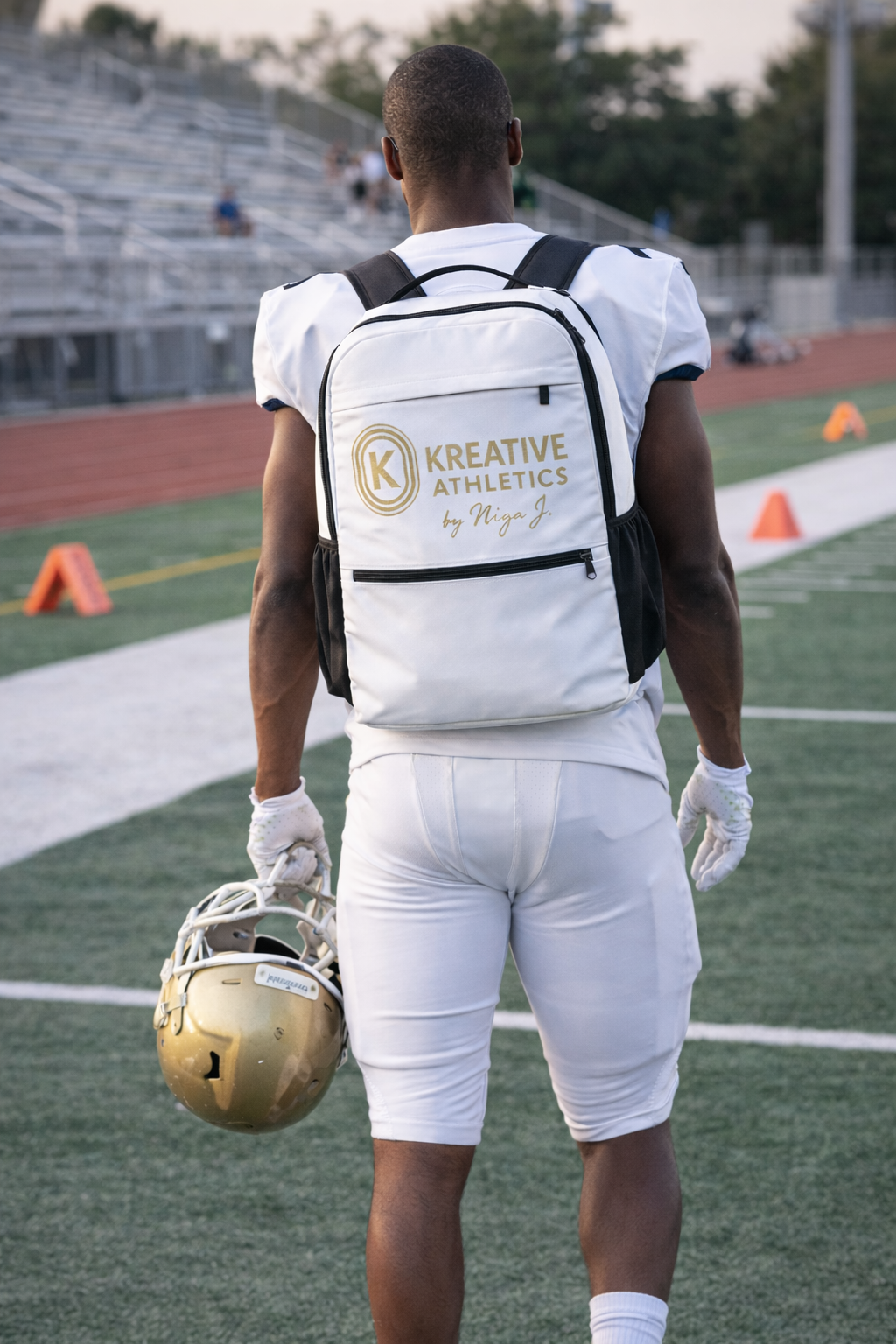 A football player stands on a field holding a helmet in his right hand, wearing a white uniform with a backpack that reads 'KREATIVE ATHLETICS by Niga J.' The stadium and track are visible in the background.