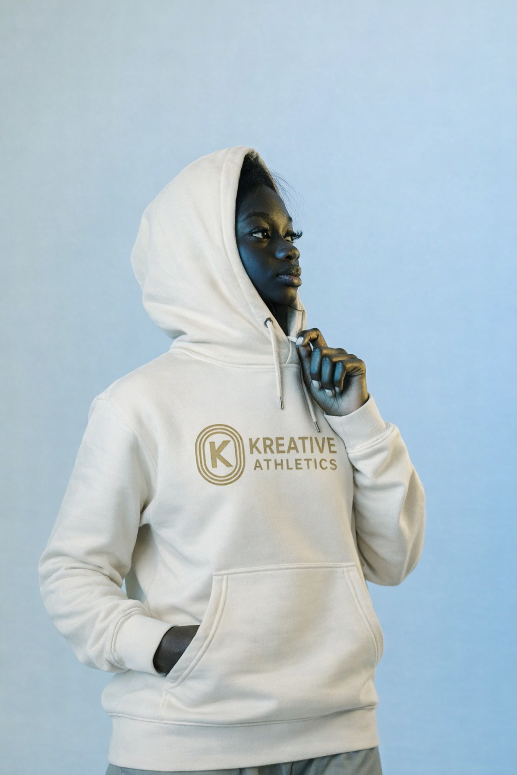 A young woman with dark skin and curly hair wearing a white hoodie with the logo "KREATIVE ATHLETICS" stands against a plain light blue background. She is looking to her left, with her right hand holding the hoodie near her chin and her left hand in 