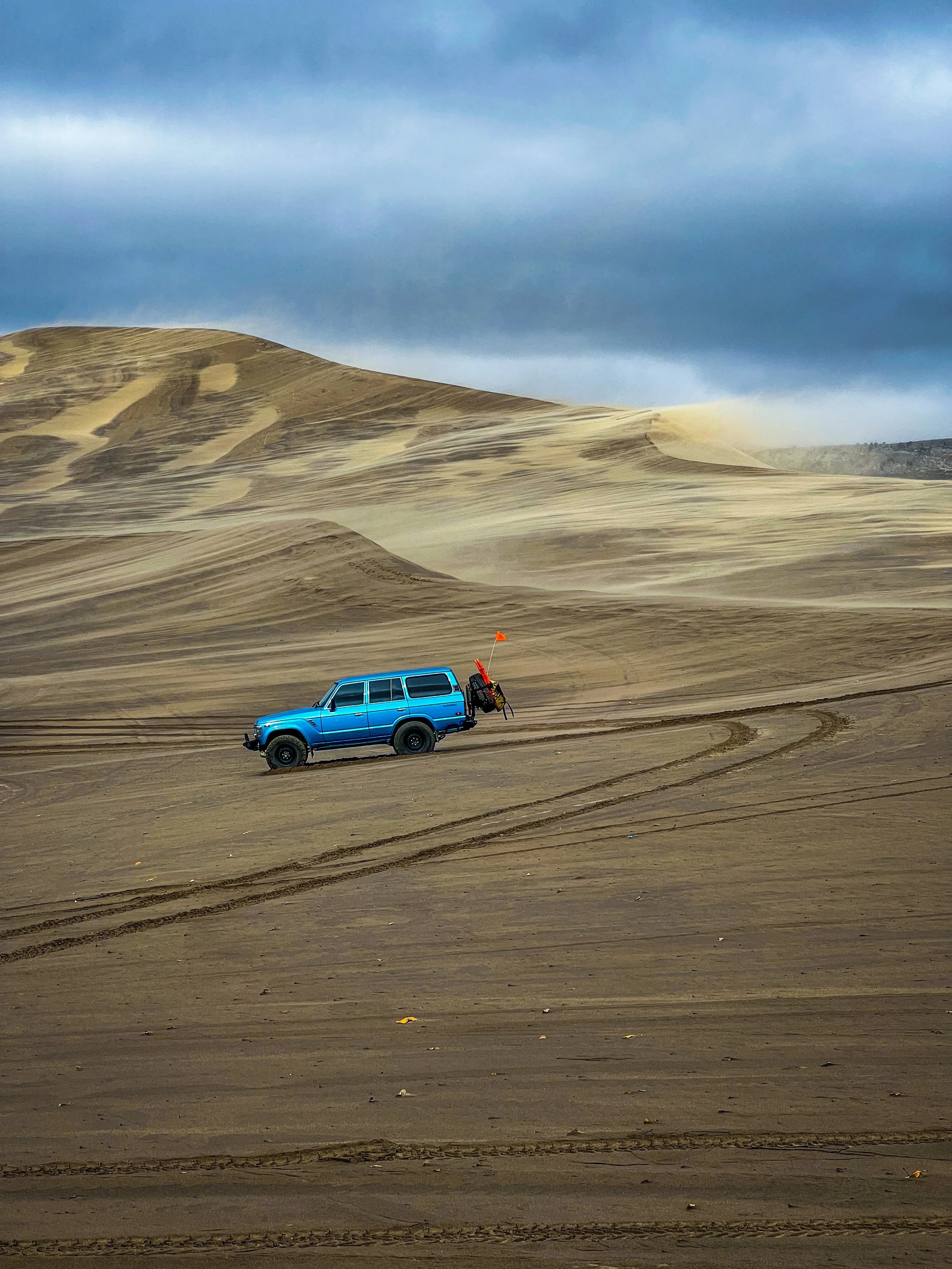 Blue SUV parked on sandy desert landscape with tire tracks and sand dunes, cloudy sky overhead.