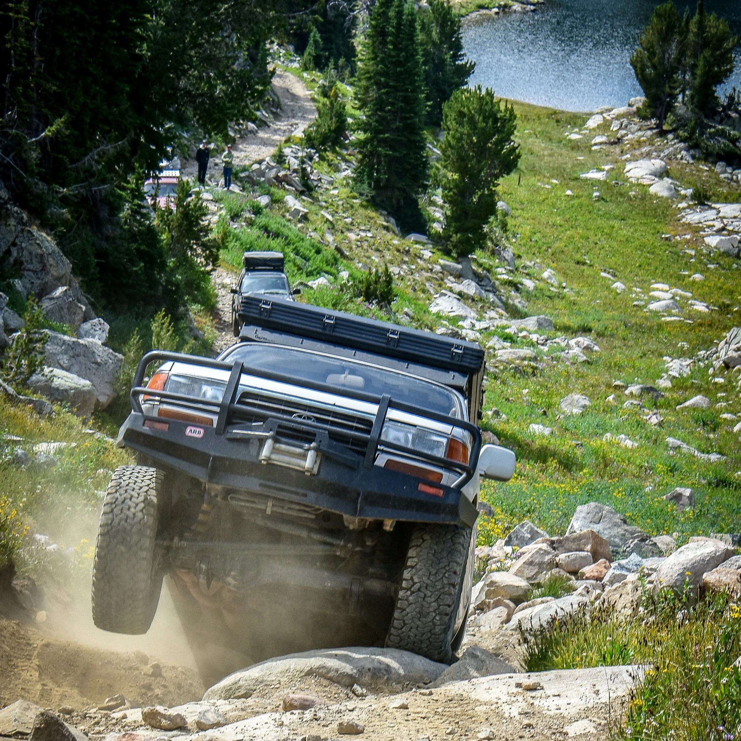 A black off-road vehicle driving on a rugged mountain trail near a lake, with two people standing on the trail surrounded by trees and rocks.