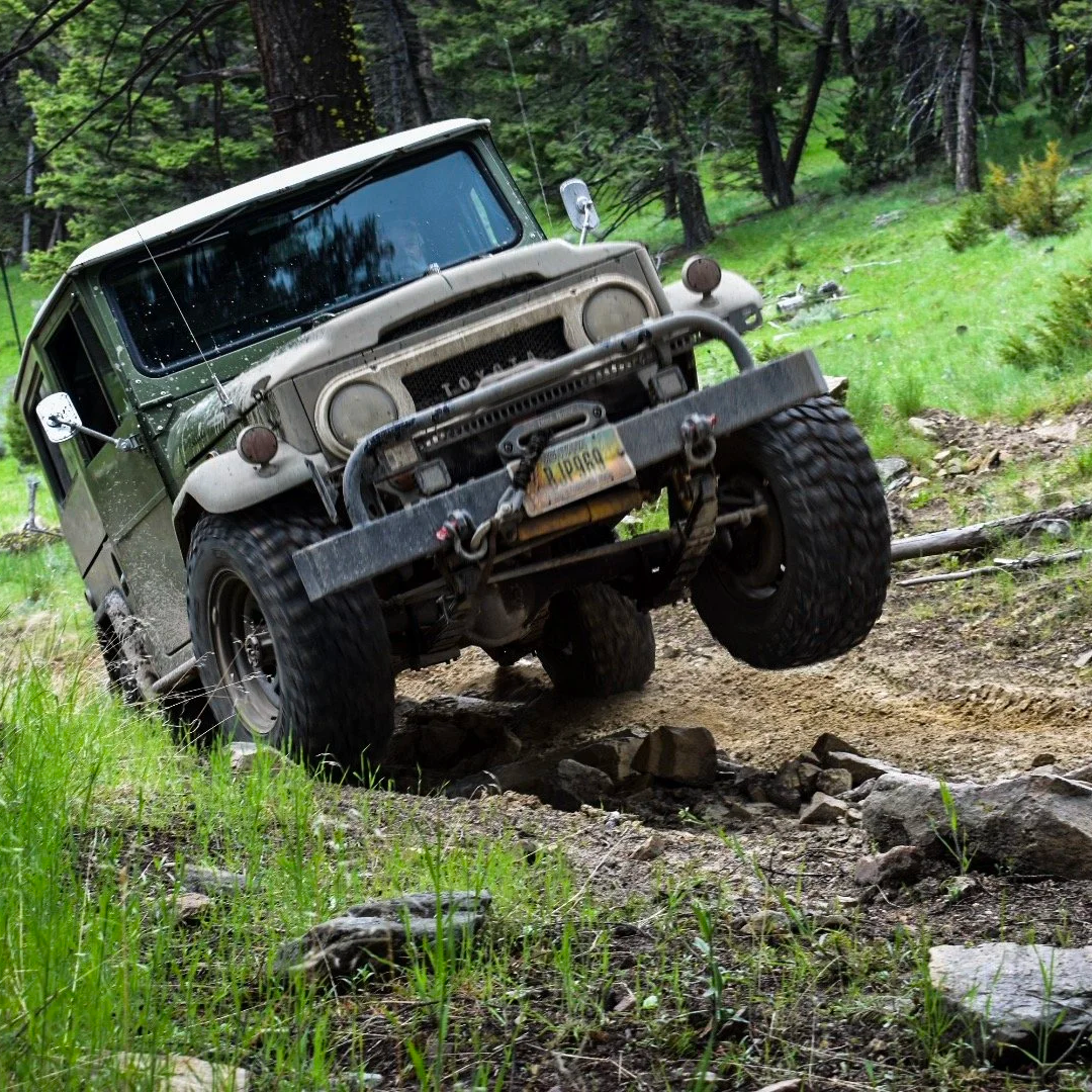 An old, rugged off-road vehicle with a green and white body, navigating rocky terrain in a forested area.