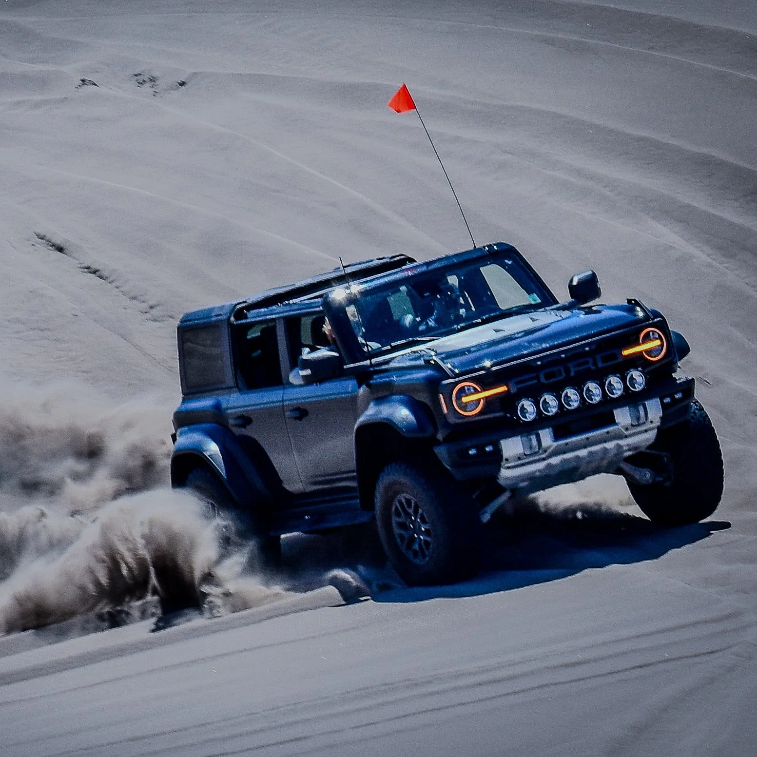 A blue Ford off-road vehicle driving on sand dunes with dust and sand kicking up behind it.
