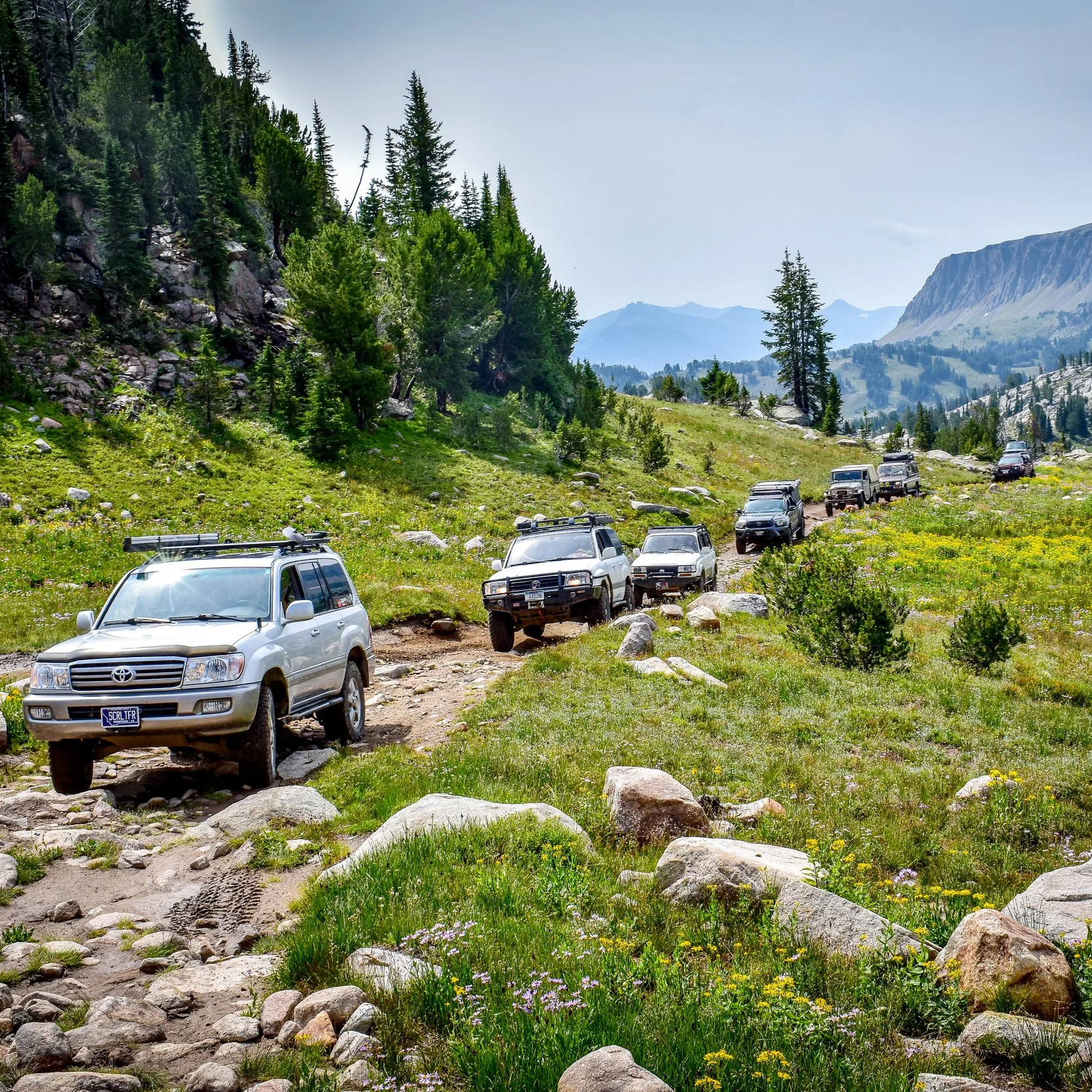 A line of off-road vehicles driving on a rugged trail through a mountainous landscape, with green grass, rocks, and trees on either side, and mountains in the background under a partly cloudy sky.