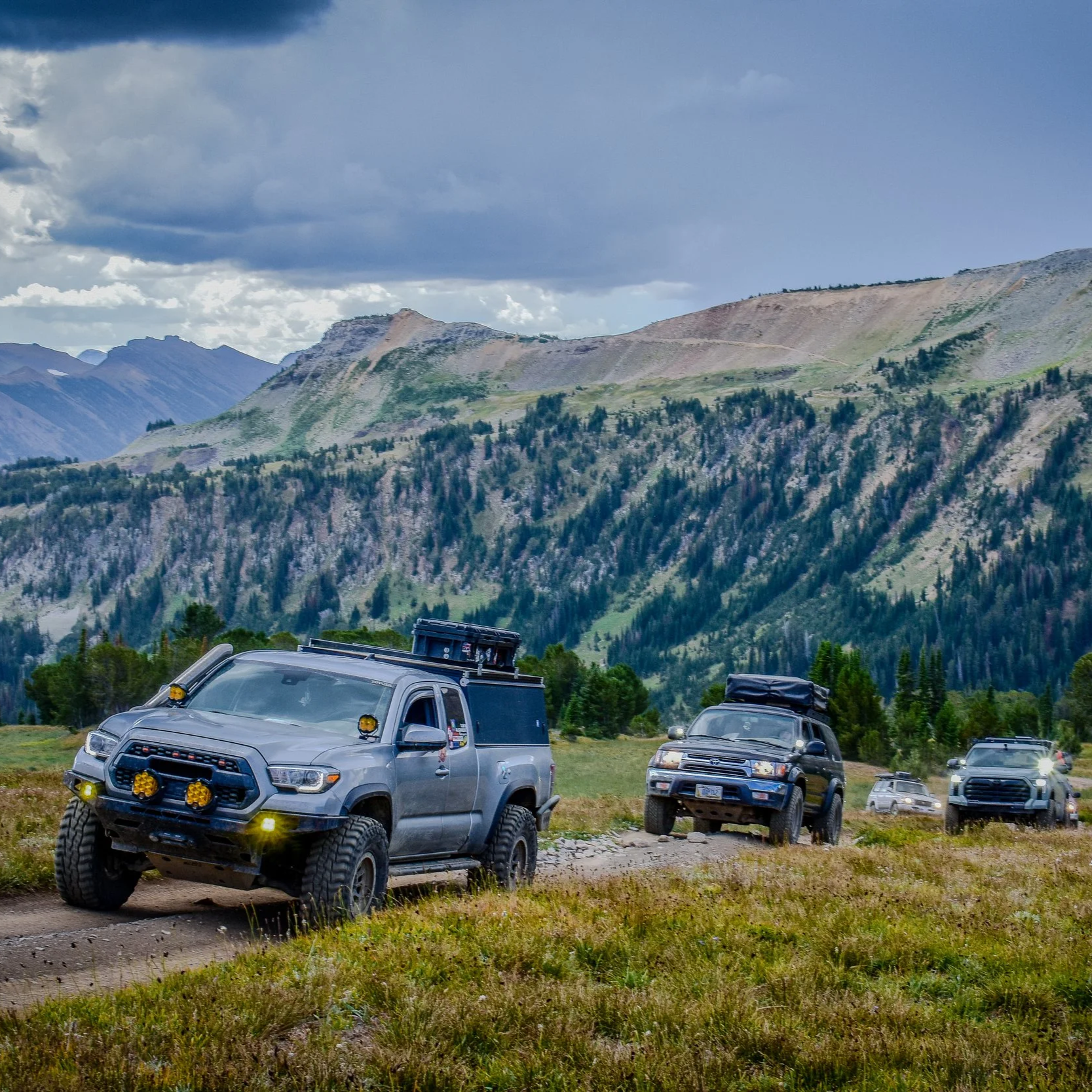A convoy of off-road vehicles driving on a dirt trail in a mountainous landscape with green hills and cloudy sky.