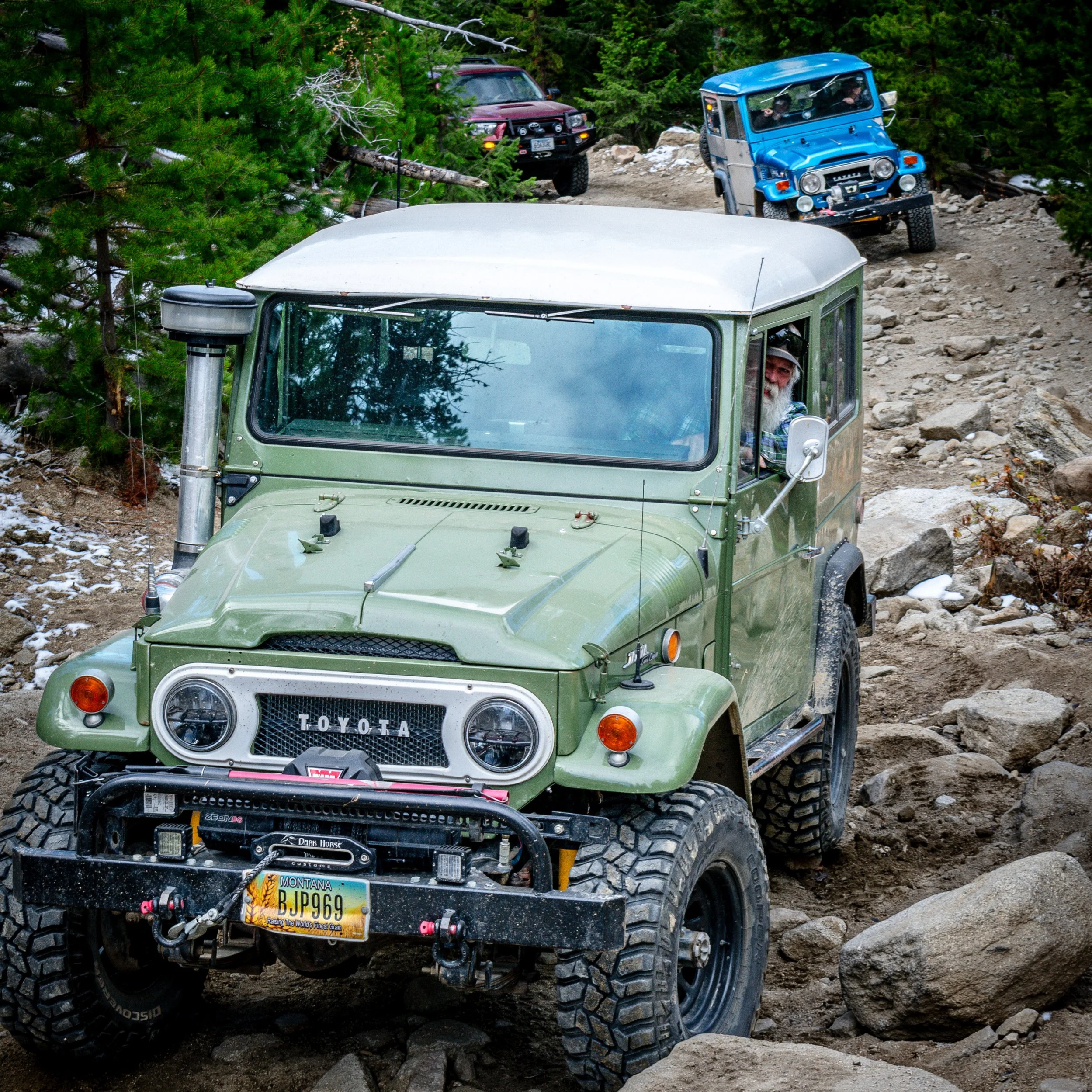 An off-road vehicle travels on a rocky trail through a forest, with two other vehicles visible in the background, one blue off-road vehicle and a dark SUV, along with a person driving the main vehicle.