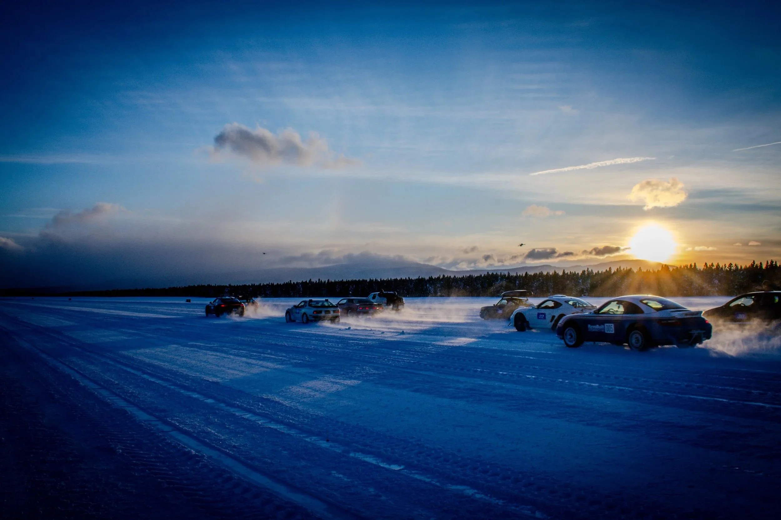 Multiple cars racing on a snowy track at sunset, kicking up snow as they speed along.