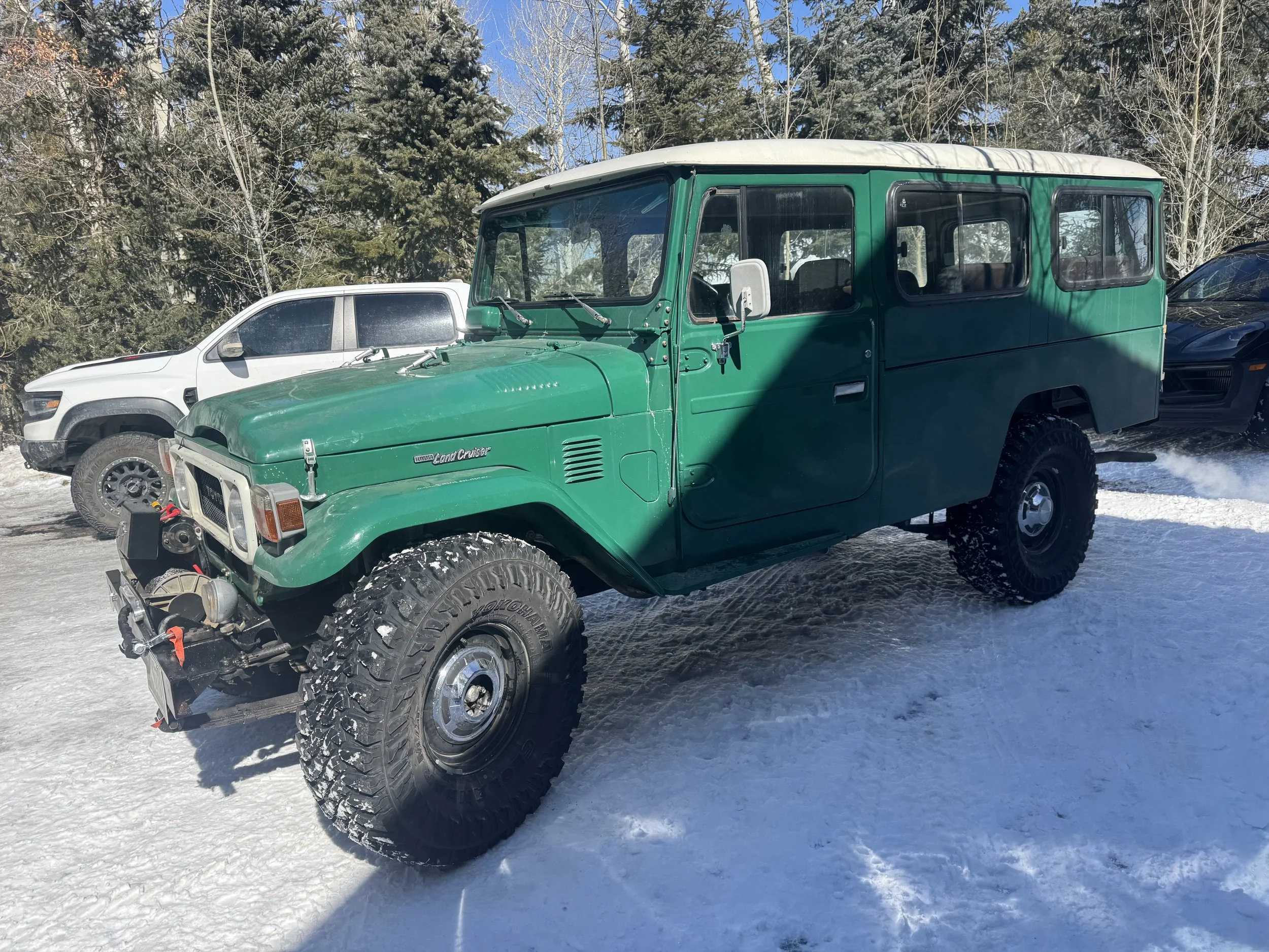 A green vintage Land Cruiser parked on a snowy surface with a white modern vehicle and trees in the background.