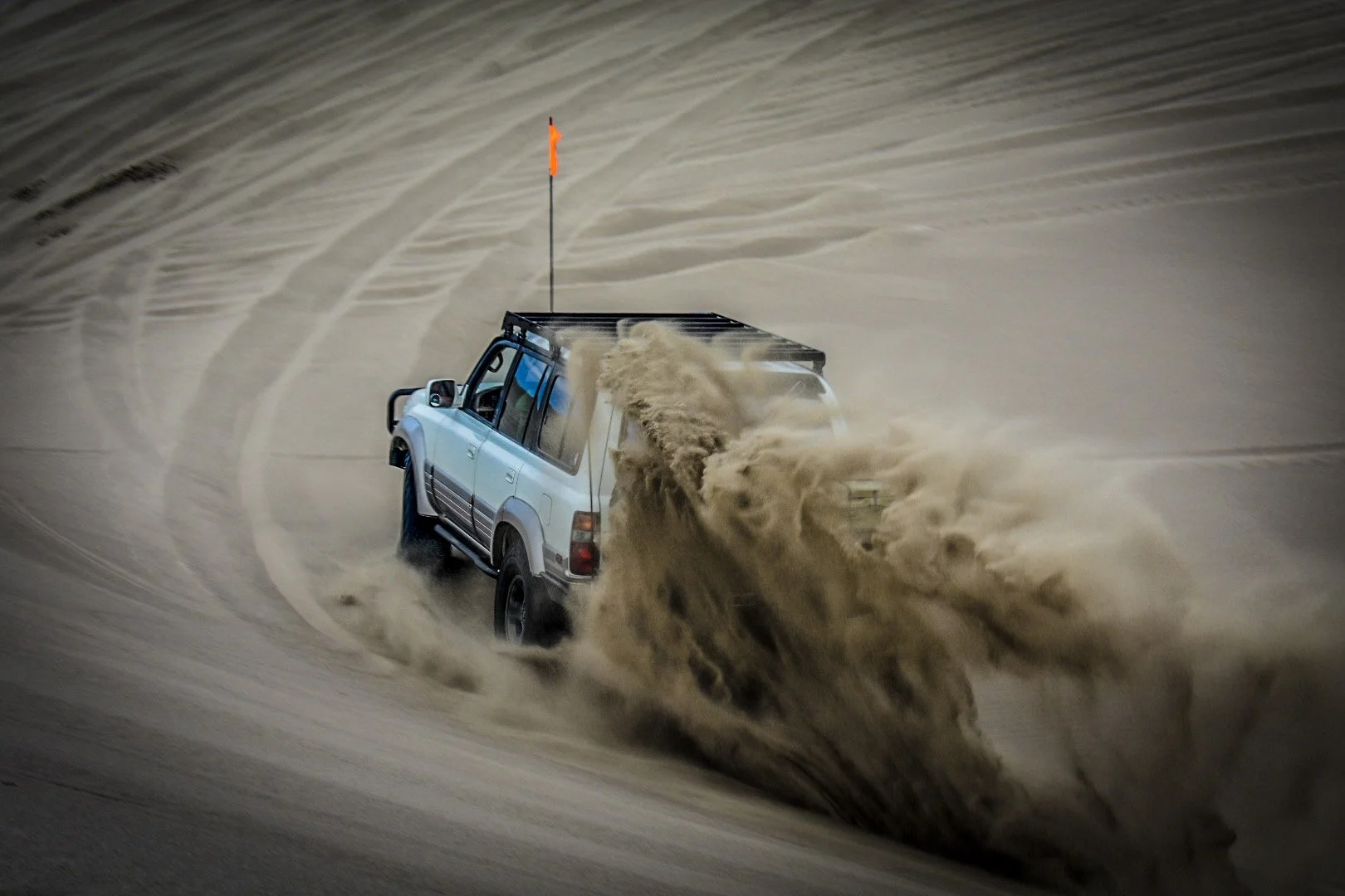 An off-road vehicle driving quickly through a sandy desert, kicking up a large cloud of sand behind it. The vehicle has a flag on a pole attached to its roof.