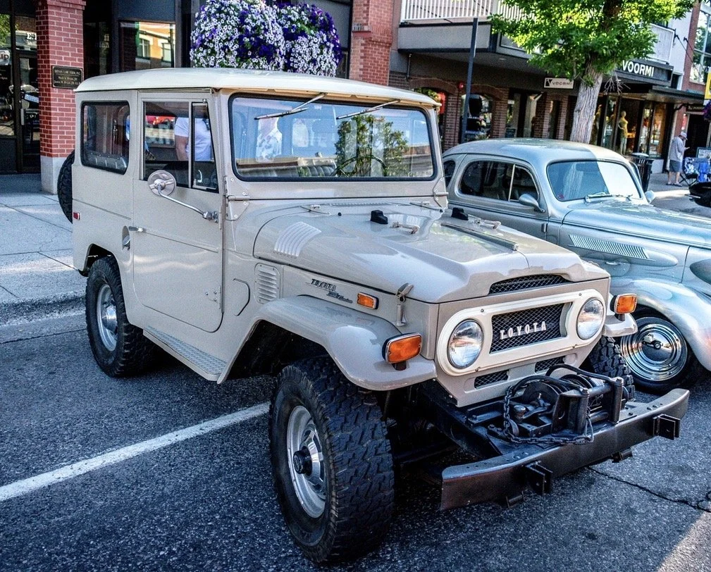 A vintage white Toyota Land Cruiser parked on the street next to a classic silver car. The Land Cruiser has a spare tire mounted on the back, a winch on the front bumper, and a large flower arrangement on its roof.