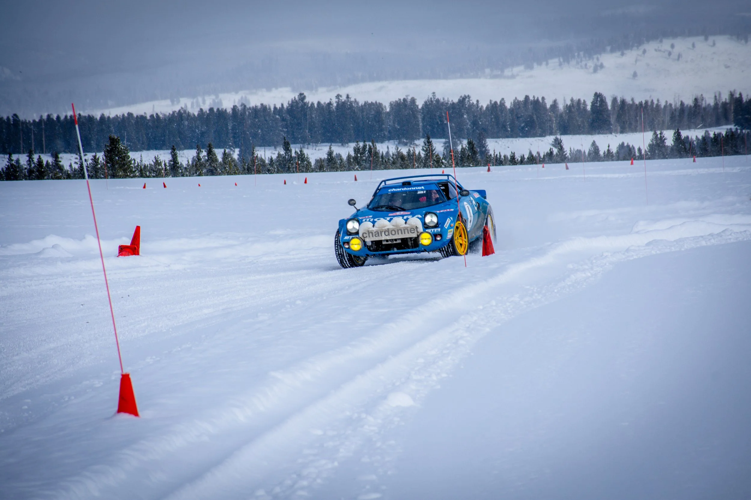 A blue vintage race car with yellow wheels driving on a snow-covered track surrounded by orange cones and markers, with snow-covered trees and mountain in the background.