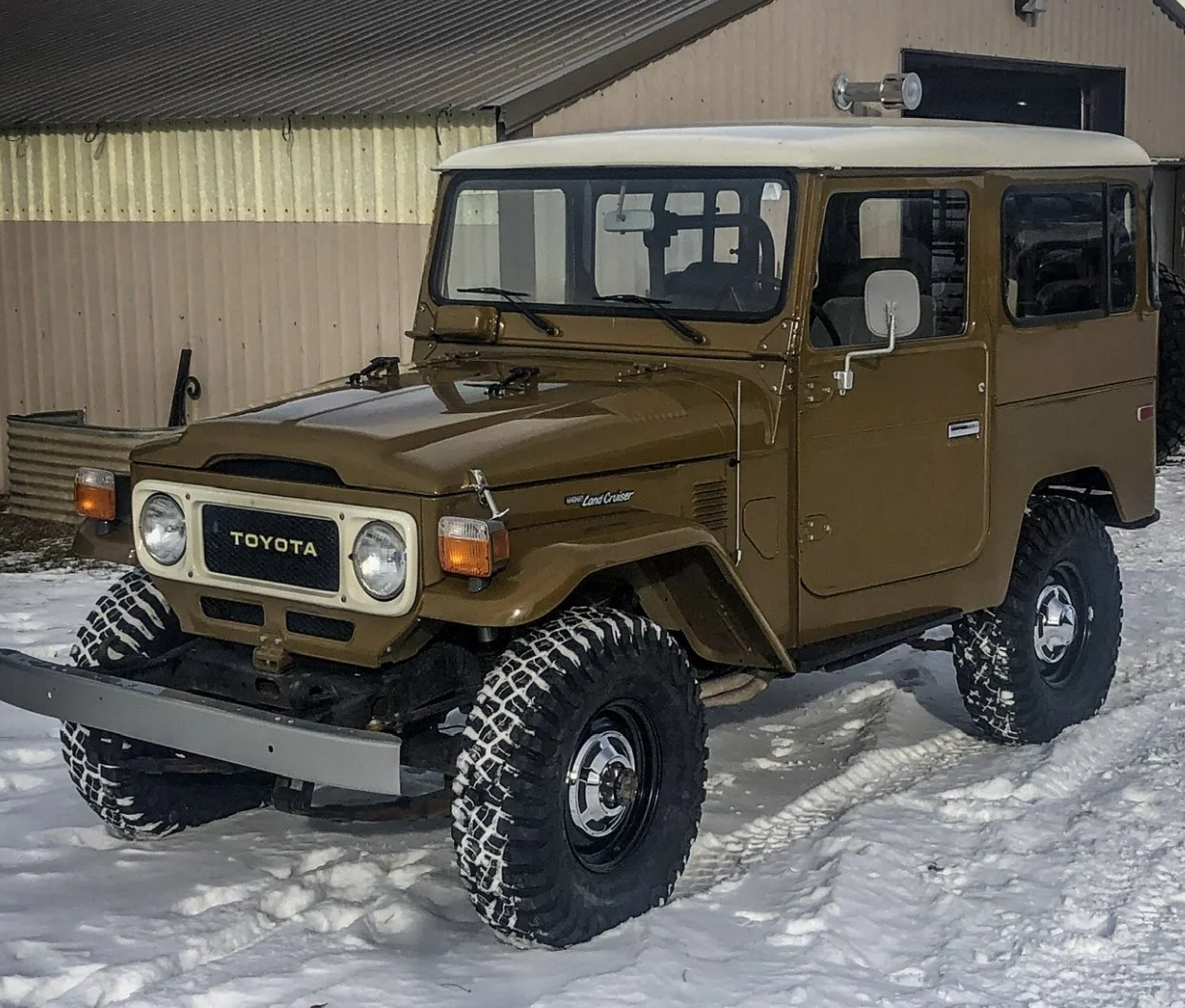 A vintage Toyota Land Cruiser parked on snow, with a beige body, white roof, large off-road tires, and a front bumper