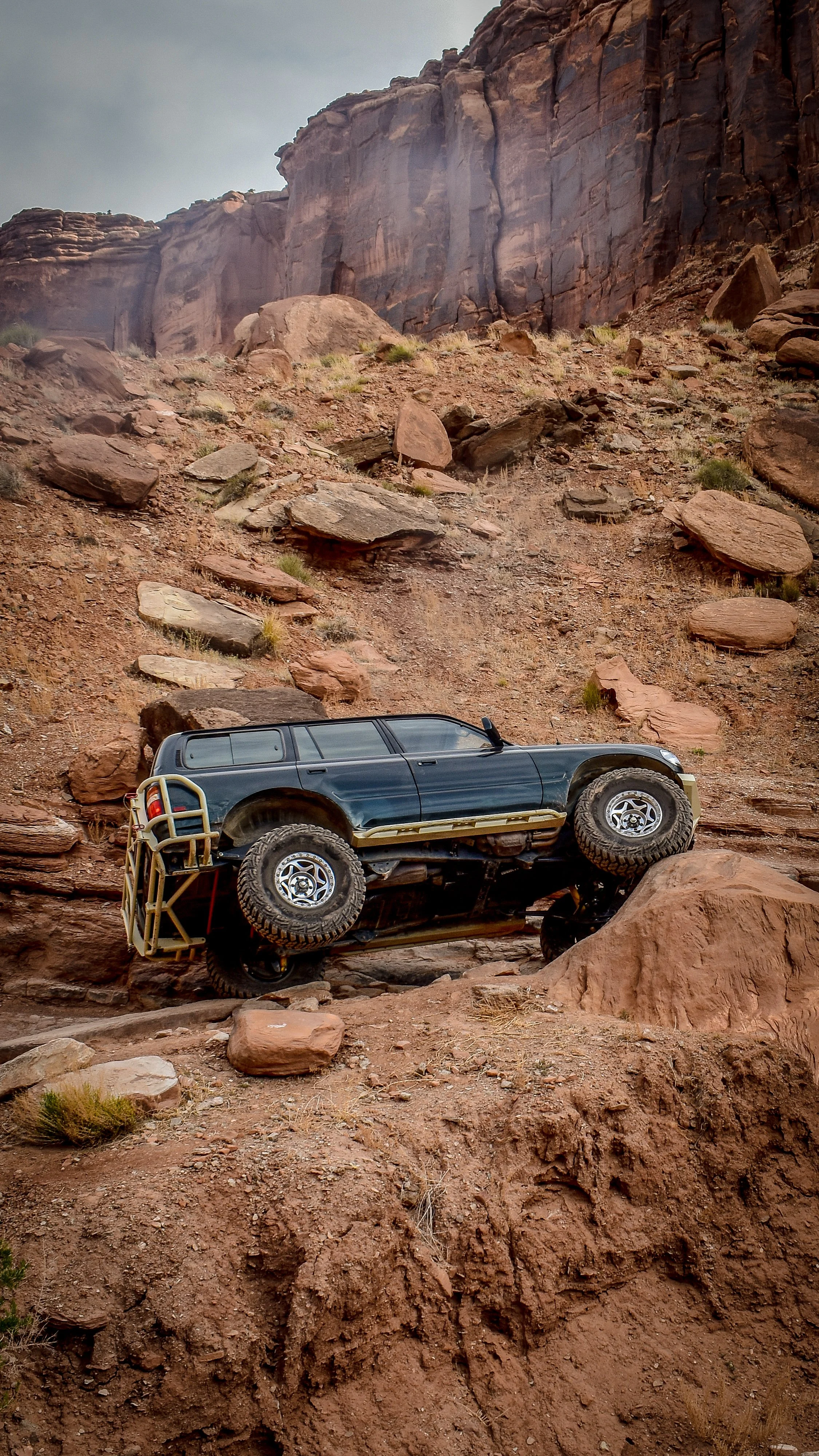 A toy off-road vehicle, resembling an SUV, positioned on rugged reddish-brown desert terrain with rocks and cliffs in the background.
