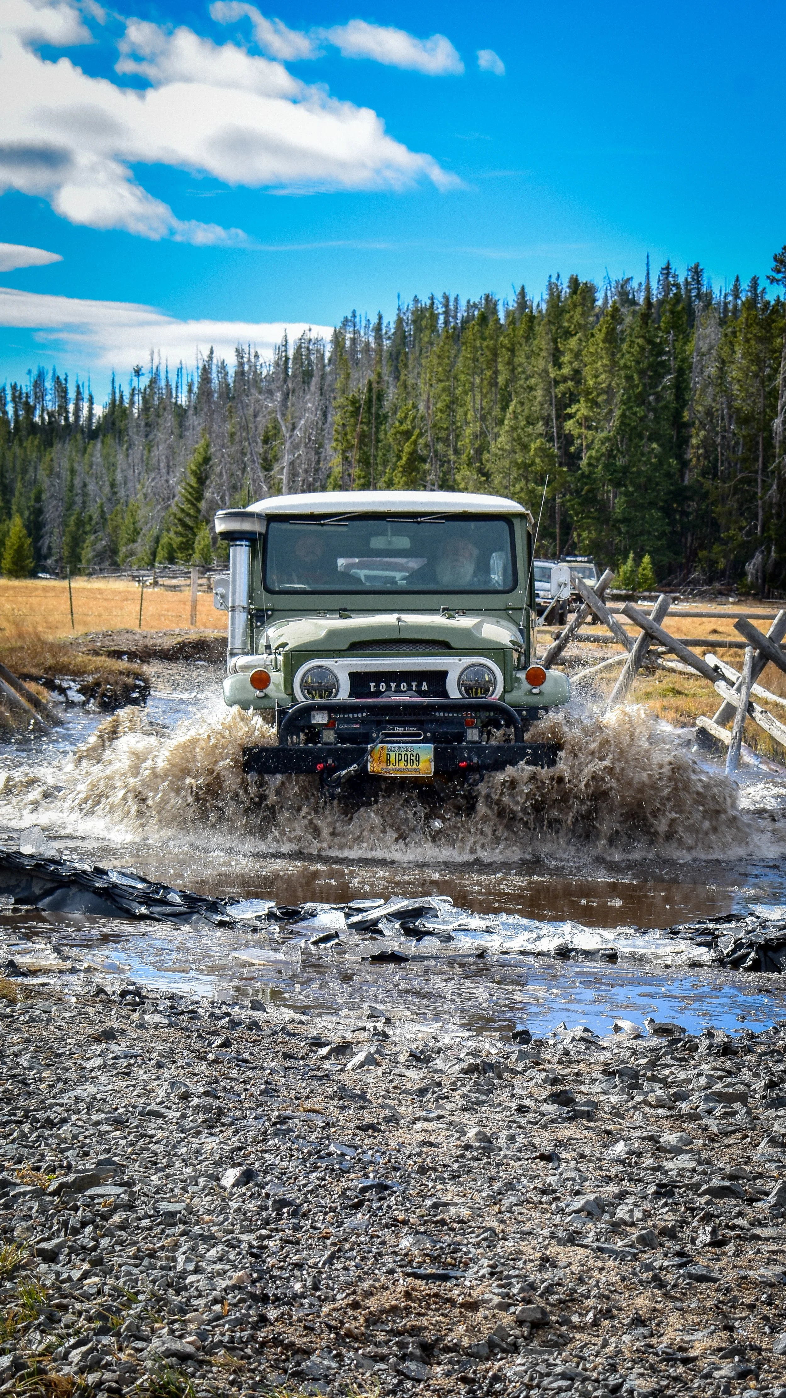 A vintage green Toyota off-road vehicle splashing through a muddy water crossing on a dirt trail in a forested area under a partly cloudy sky.