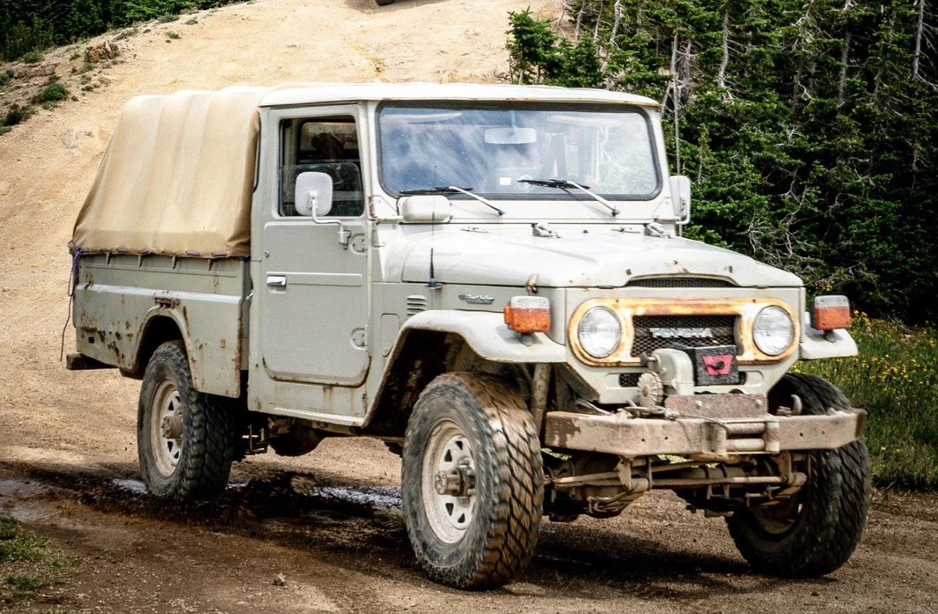 A weathered, off-road Toyota pickup truck with a beige canopy driving on a dirt path surrounded by trees and greenery.