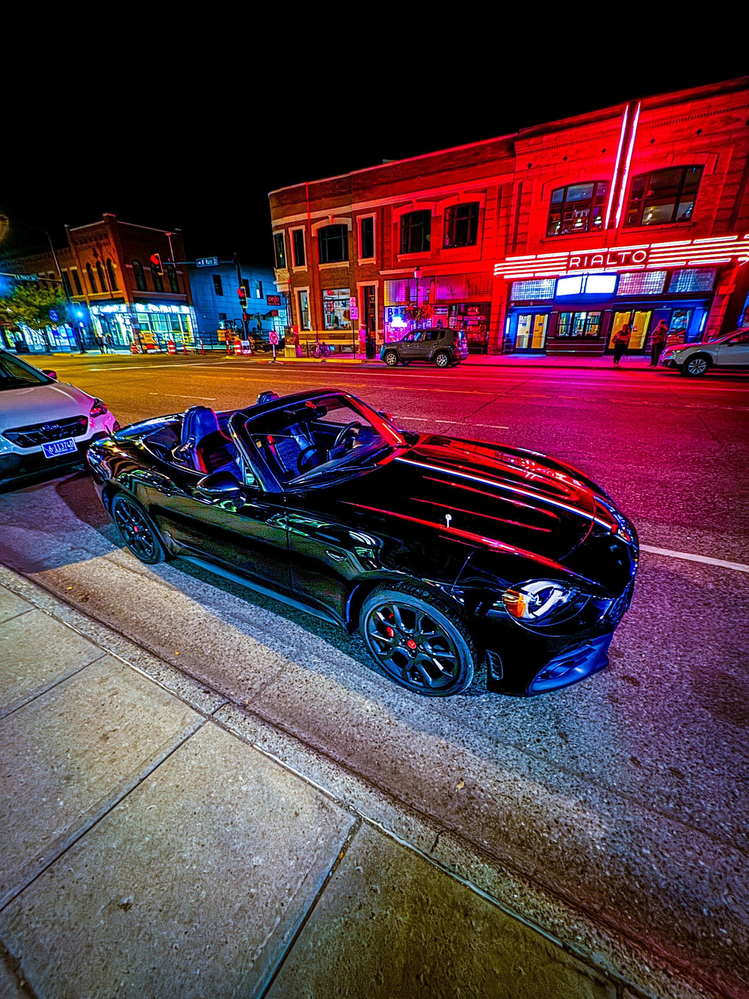 A black convertible sports car parked on a city street at night, with colorful neon lights illuminating buildings and signs in the background.